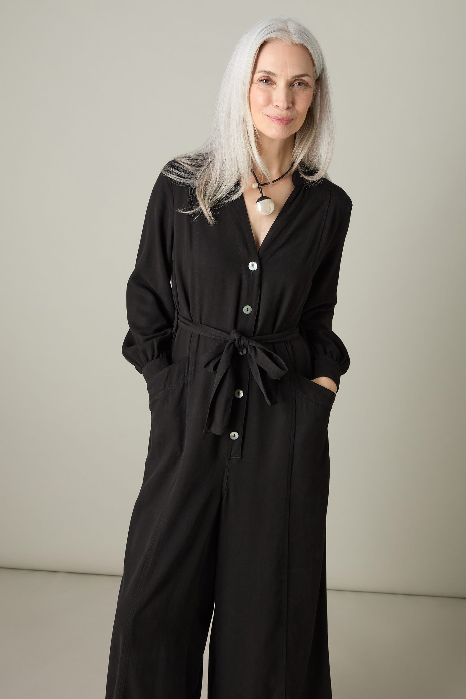 A person with long gray hair stands smiling, wearing a stylish black Fluid Twill Jumpsuit made from sustainable materials, with hands in pockets against a neutral background.