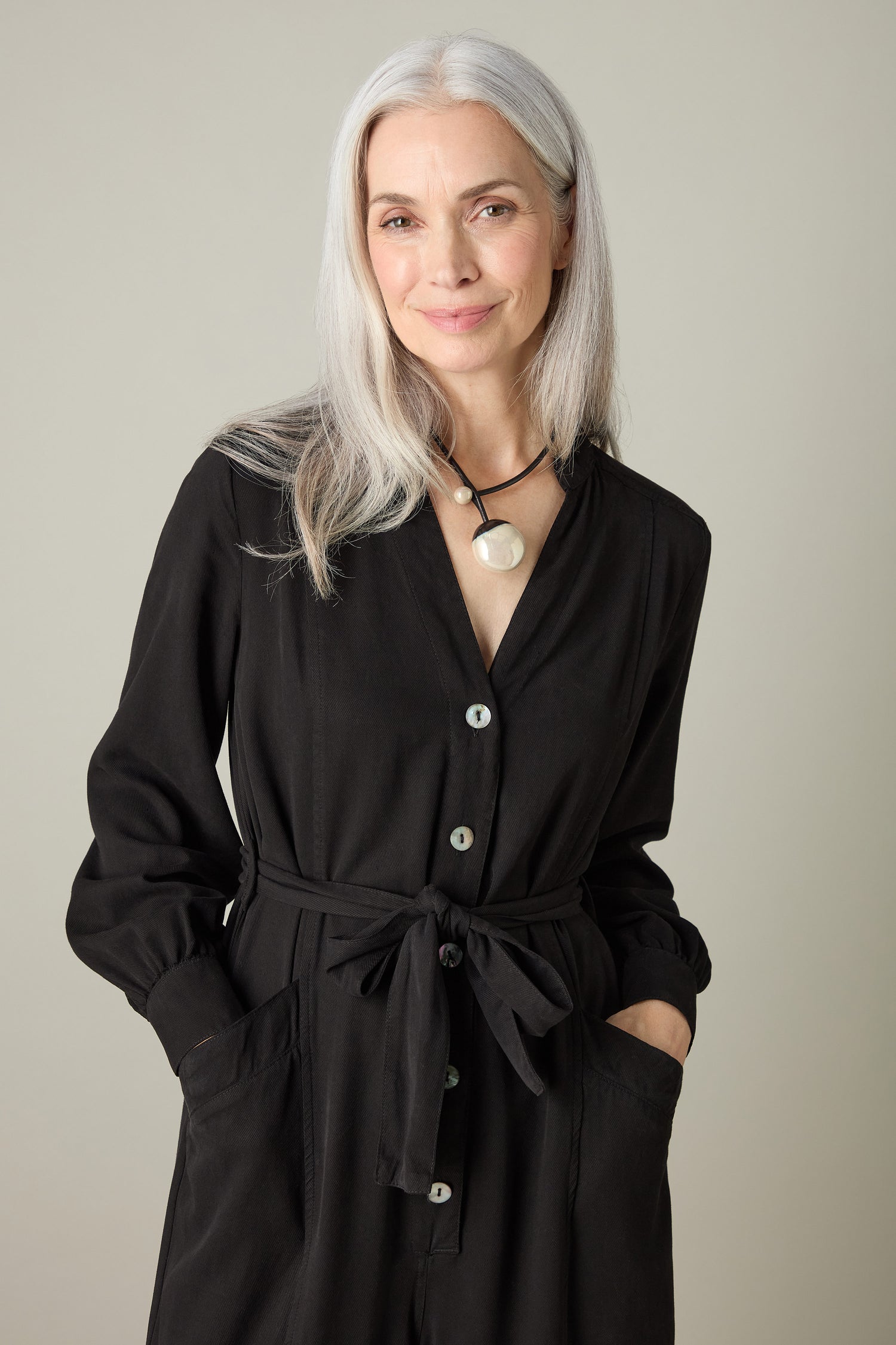 A person with long white hair stands elegantly in a black Fluid Twill Jumpsuit, crafted from eco-friendly fabric, hands tucked in pockets. A necklace with a round pendant complements the outfit as they pose gracefully against a plain background.