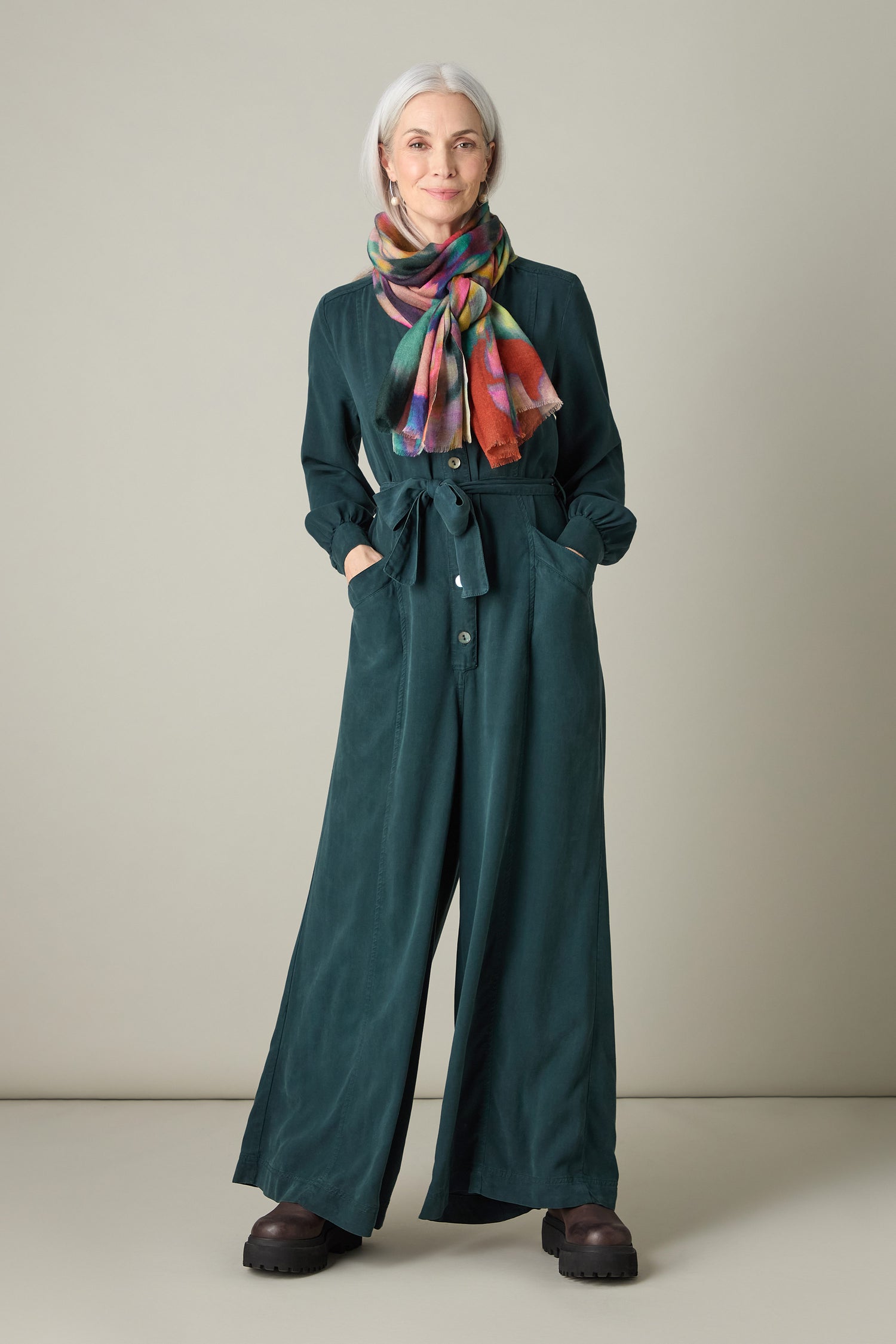 An individual sporting gray hair is dressed in the eco-friendly Fluid Twill Jumpsuit, which showcases a sleek rounded neckline. Their hands are casually placed in the pockets, and they complement the outfit with brown shoes and a vibrant scarf against an unadorned background.