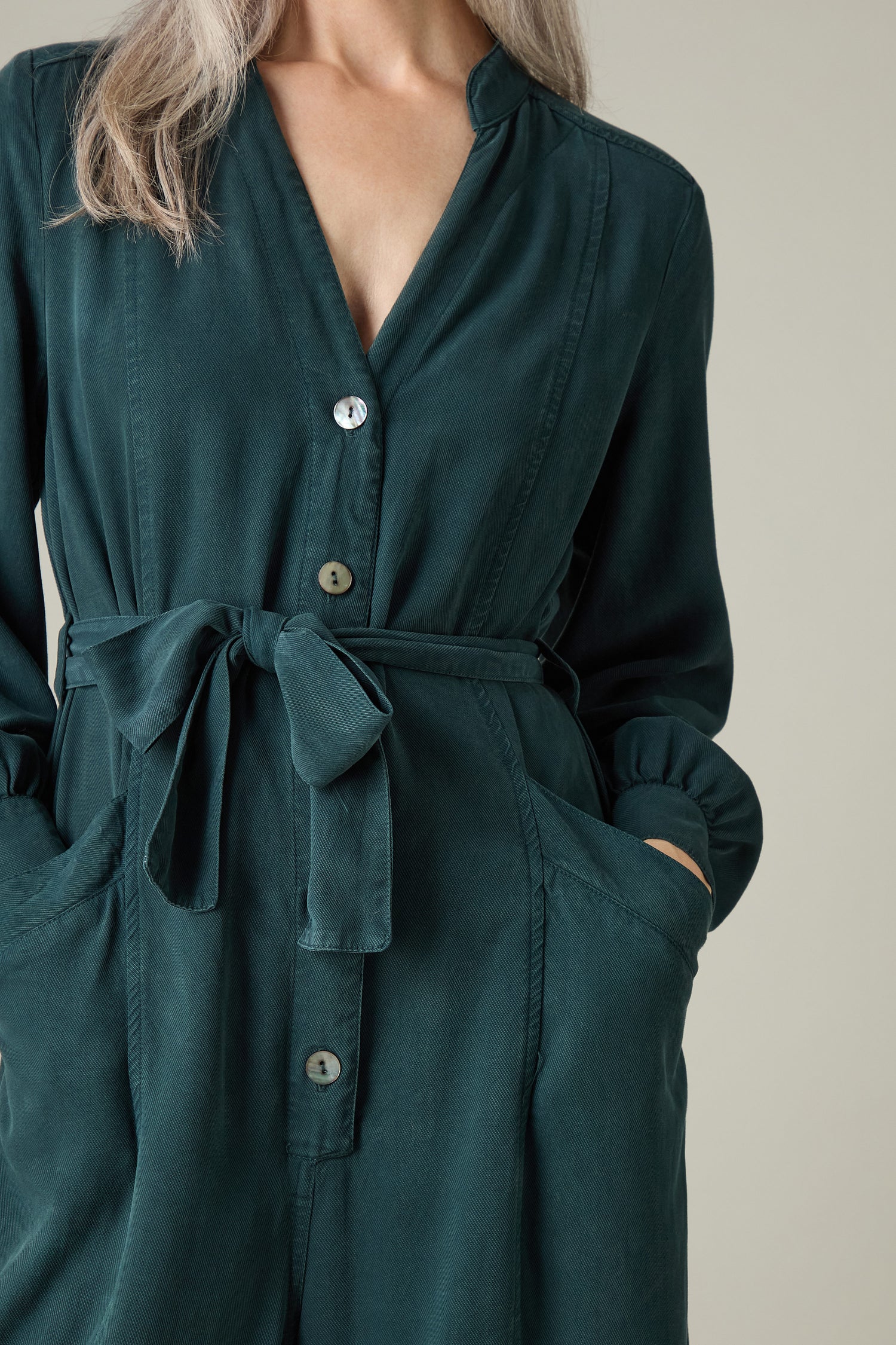 A person dressed in a Fluid Twill Jumpsuit in a deep green shade, featuring eco-friendly material, a tied waist sash, and hands casually placed in pockets.