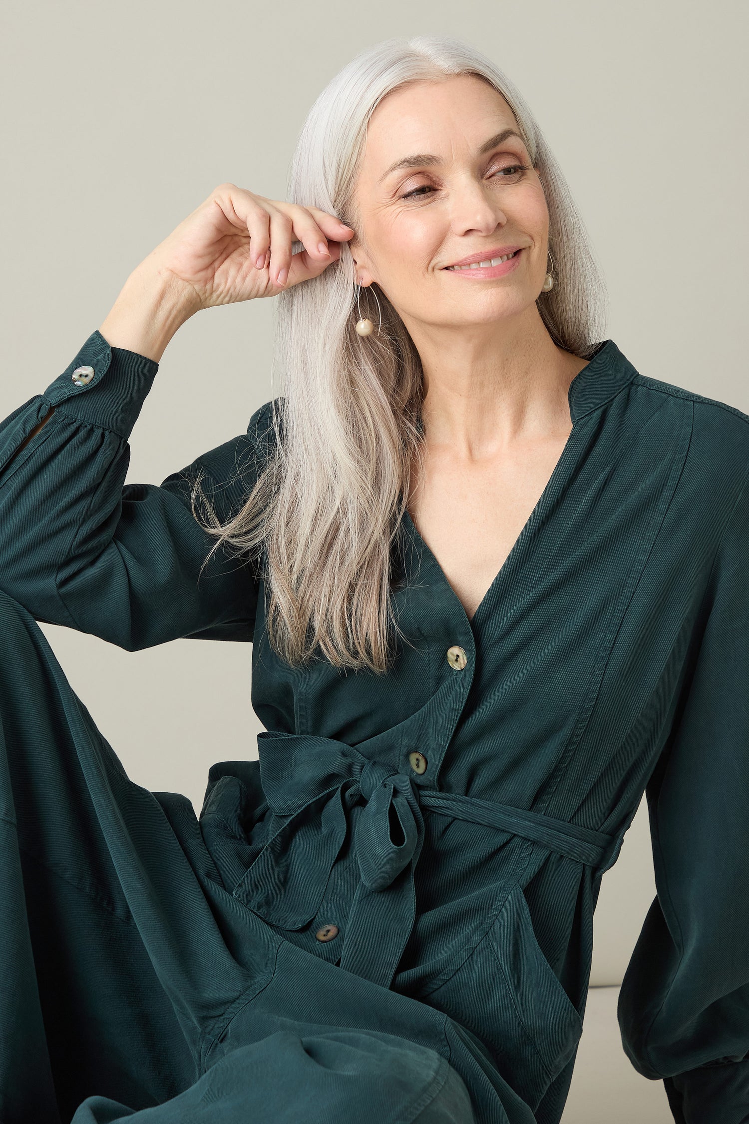 A woman with long gray hair, wearing a stylish dark green Fluid Twill Jumpsuit made from eco-friendly fabric, smiles while seated in front of a plain background.