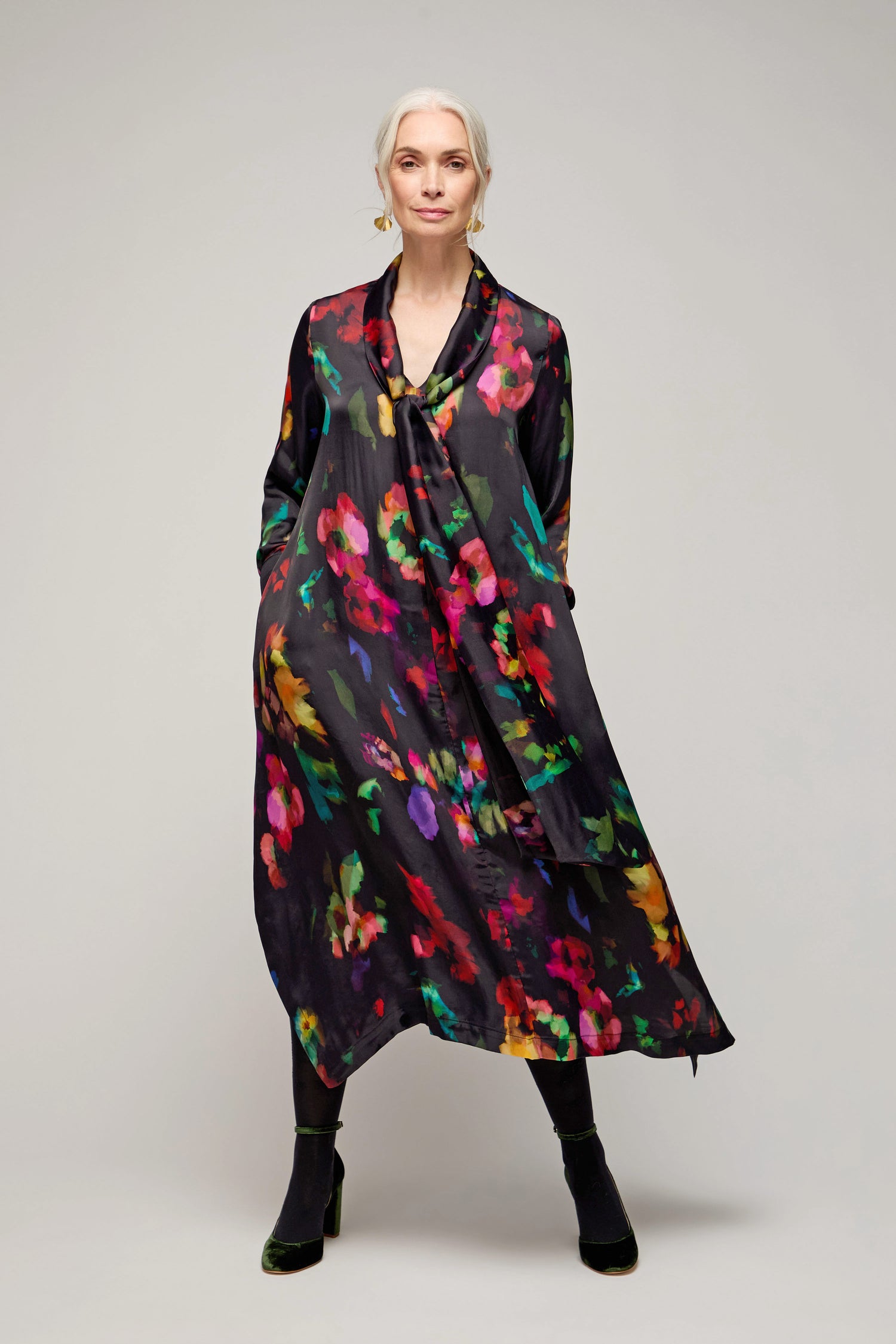 A woman with grey hair stands with hands in pockets, wearing the Winter Floral Tie Neck Dress with a vibrant abstract floral print and black boots, set against a plain light background.