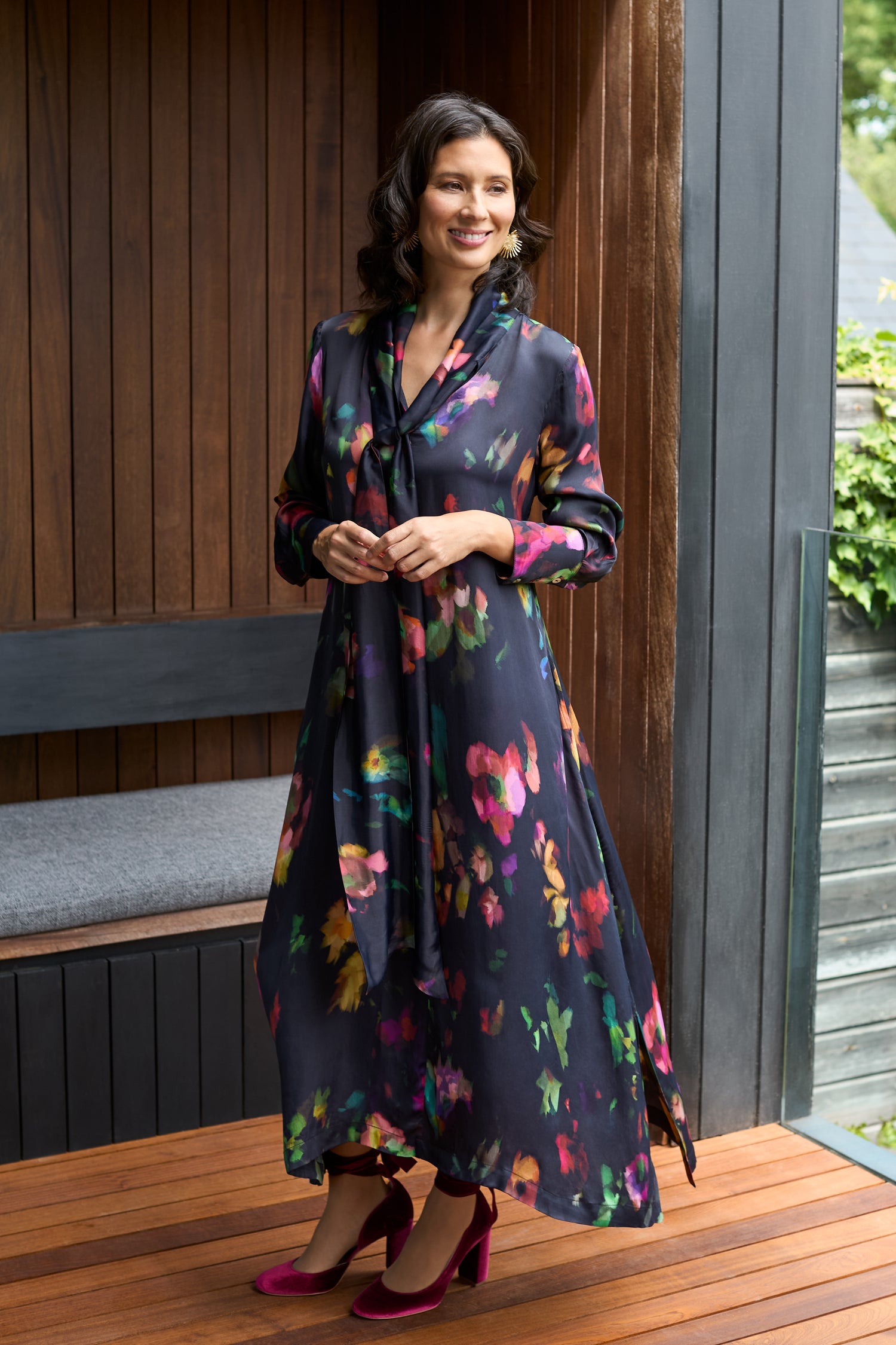 A woman smiles and looks to the side while standing on a wooden porch, wearing the Winter Floral Tie Neck Dress with a relaxed fit and red heels.