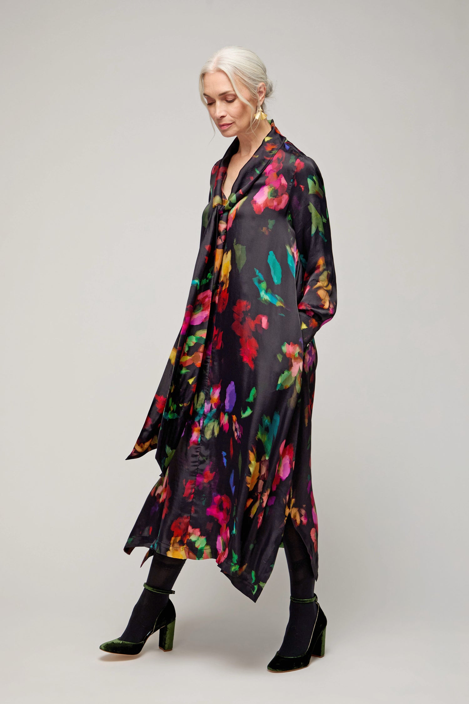 A woman with gray hair wears the Winter Floral Tie Neck Dress, black tights, and black velvet shoes while standing against a plain light background.