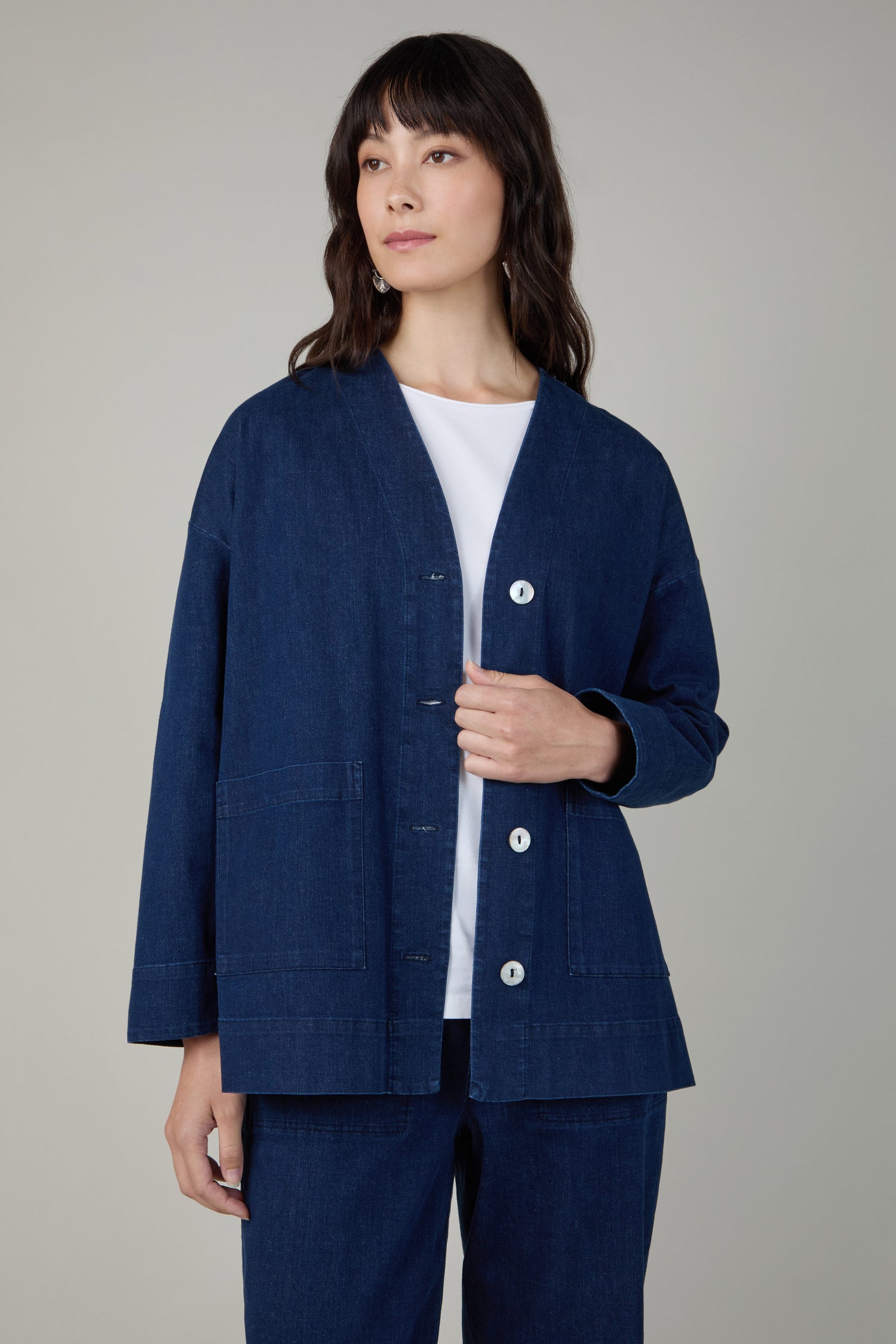A person stands against a plain background wearing the Stretch Denim Boxy Jacket in dark blue with white buttons, paired with a white top and matching pants.