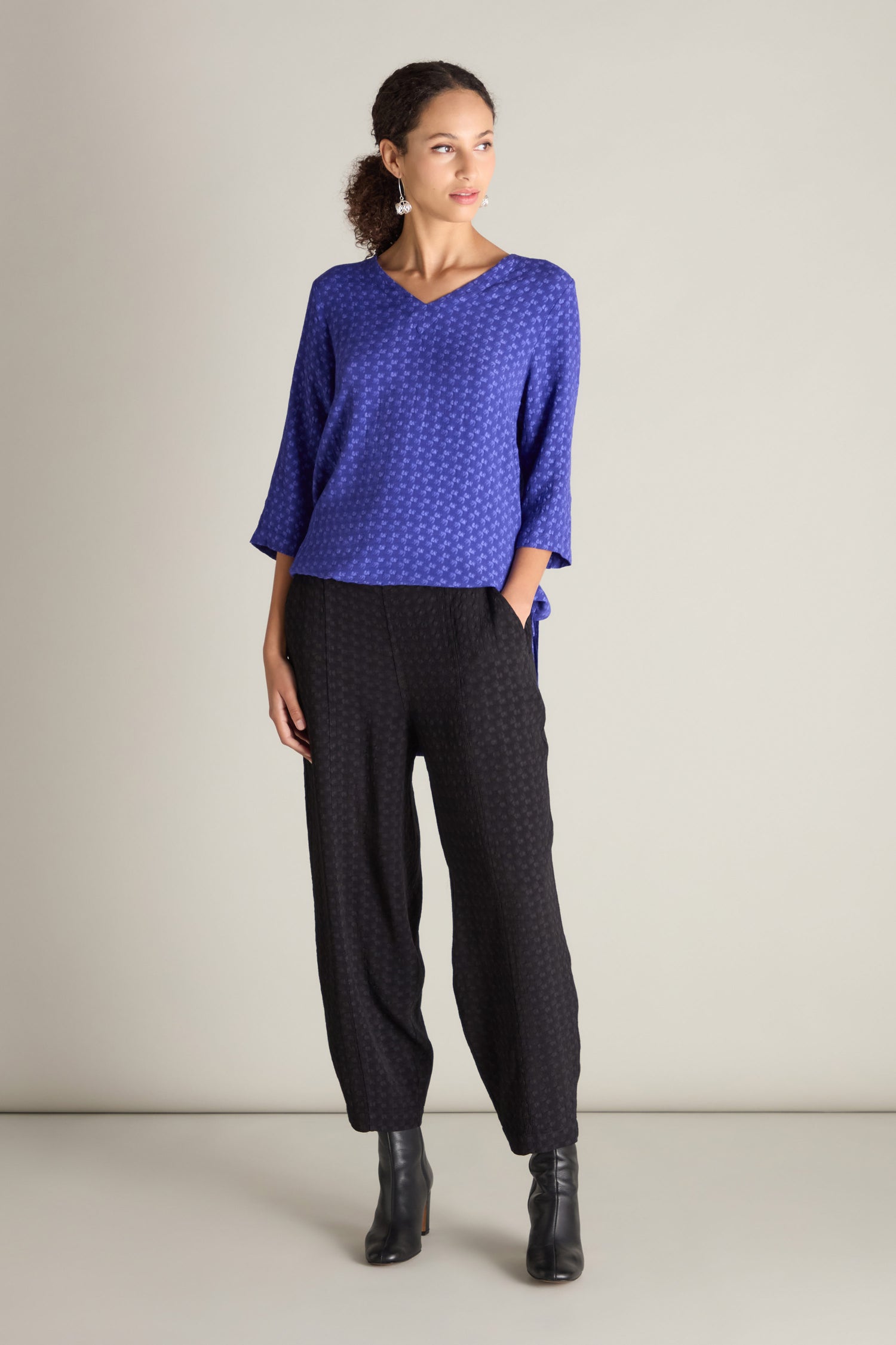 A woman donning a blue patterned top, chic silhouette black Small Square Jacquard Bubble Trousers with an elasticated waistband, and sleek black boots is standing with one hand in her pocket, gazing to the side.