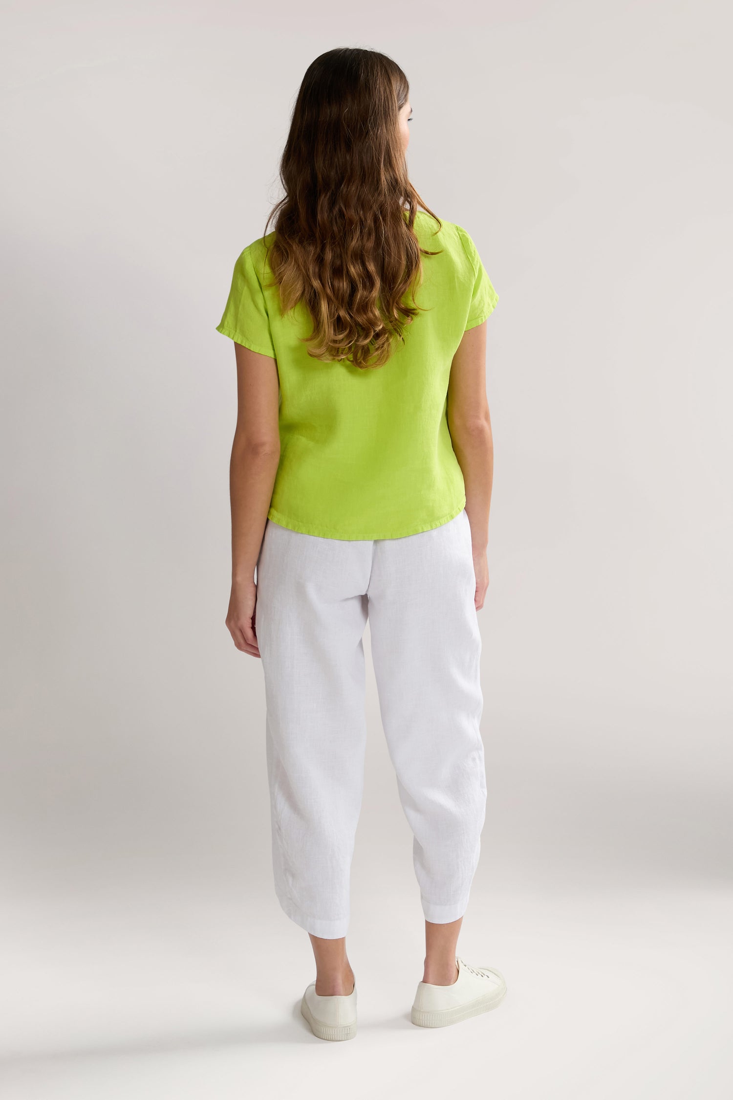 A woman with long wavy hair stands facing away, wearing a bright green short-sleeved shirt, Linen Bubble Trouser, and white sneakers against a simple, light-colored background.