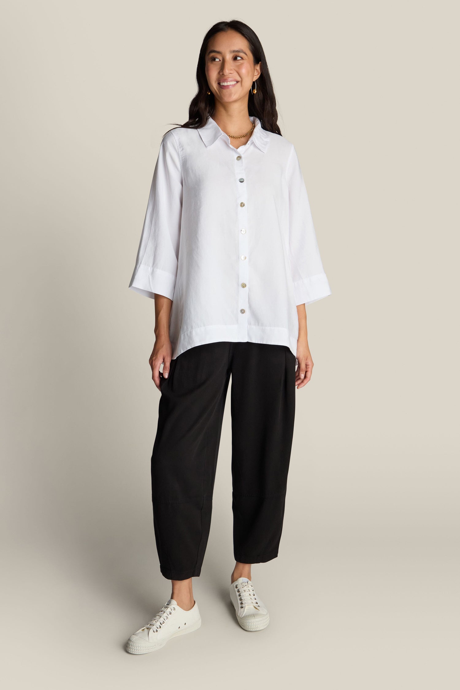 A woman stands smiling, wearing a white button-up shirt with three-quarter sleeves and the black Fluid Twill Bubble Trouser featuring an elasticated waist, paired with white sneakers. She has long dark hair and stands against a plain beige background.