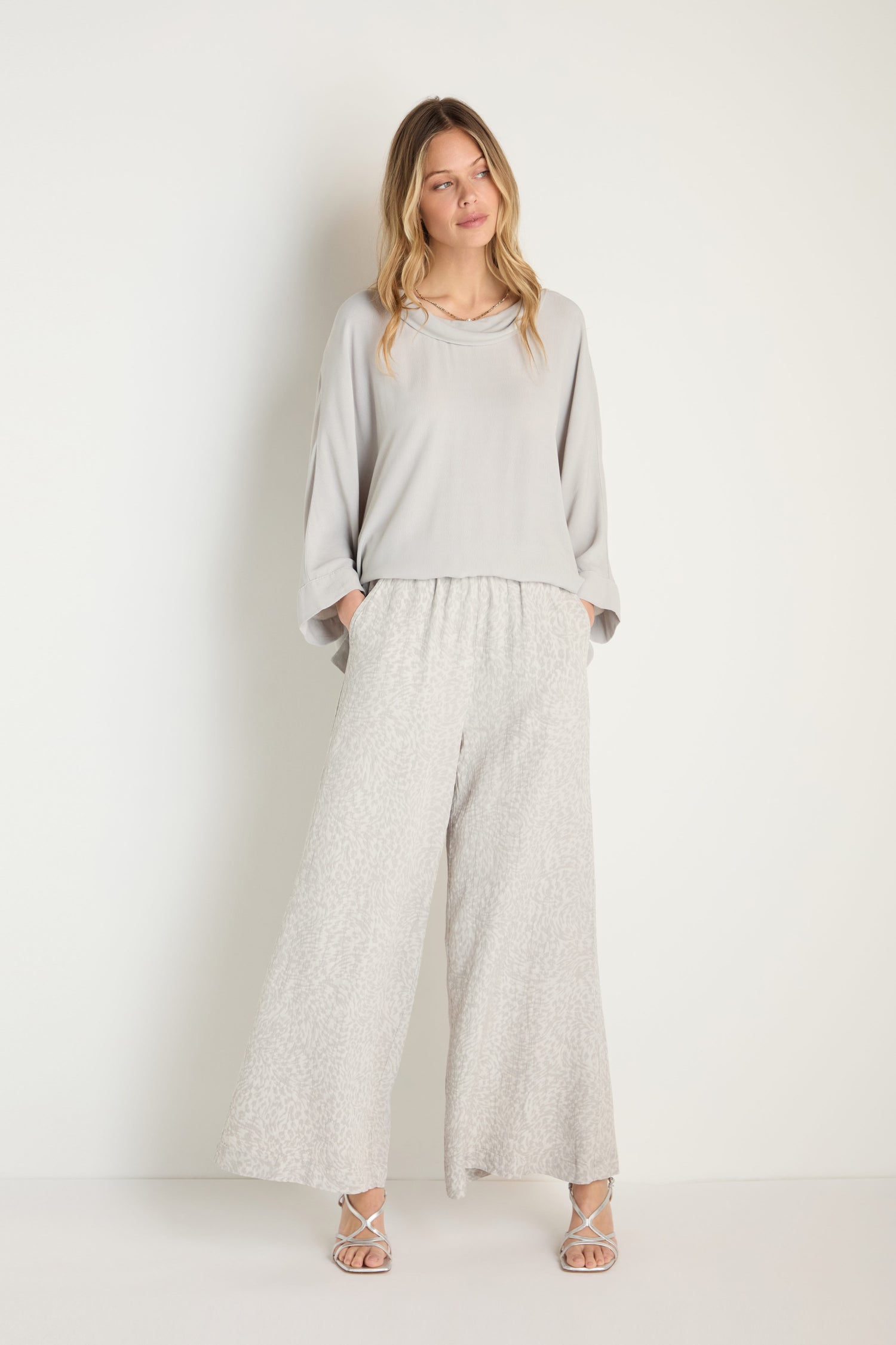 A woman stands against a white background wearing a loose light grey long-sleeve top, Animal Textured Jacquard Wide Trouser (MTP6715-ATJ), and strappy sandals, with her hands casually tucked in her pockets.