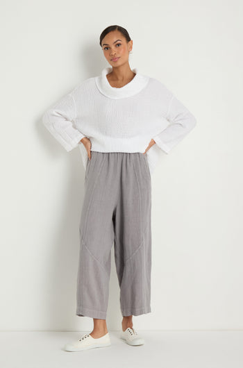 A woman stands against a white wall wearing a loose white sweater, Linen Curved Seam Bubble Trouser in gray, and white sneakers, with her hands on her hips.