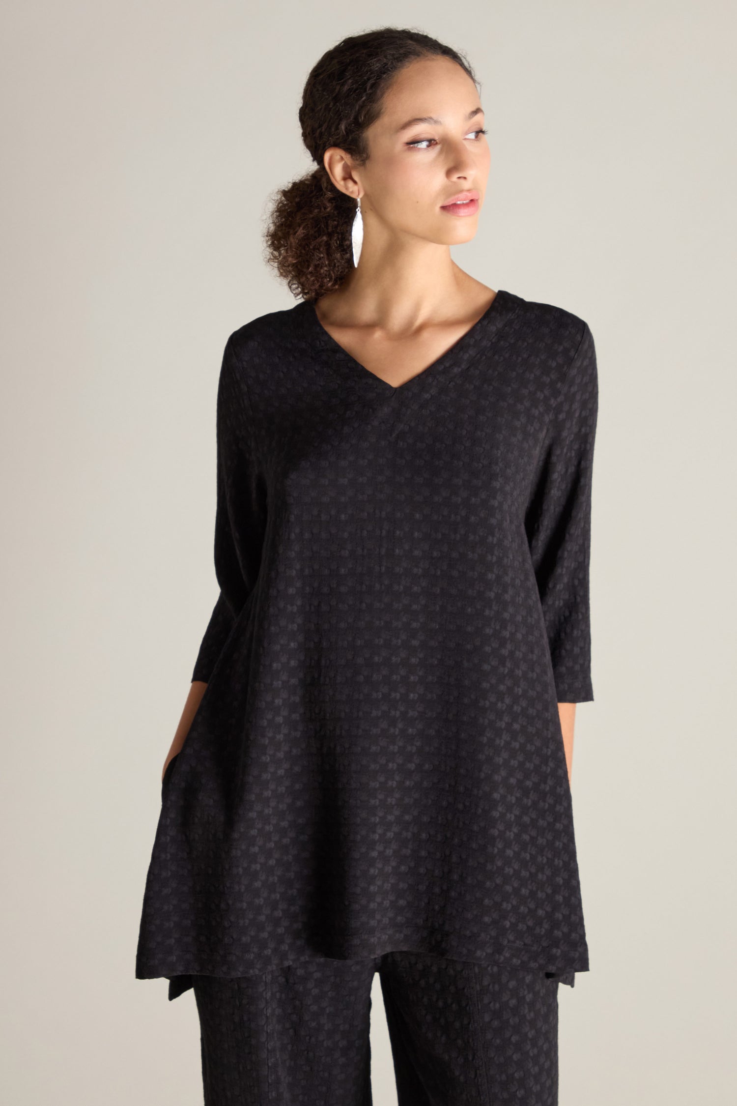 A person is wearing a black, textured Small Square Jacquard Tunic with a V-neckline and 3/4 length sleeves, paired with matching pants. They are looking to the right against a plain, light-colored background.