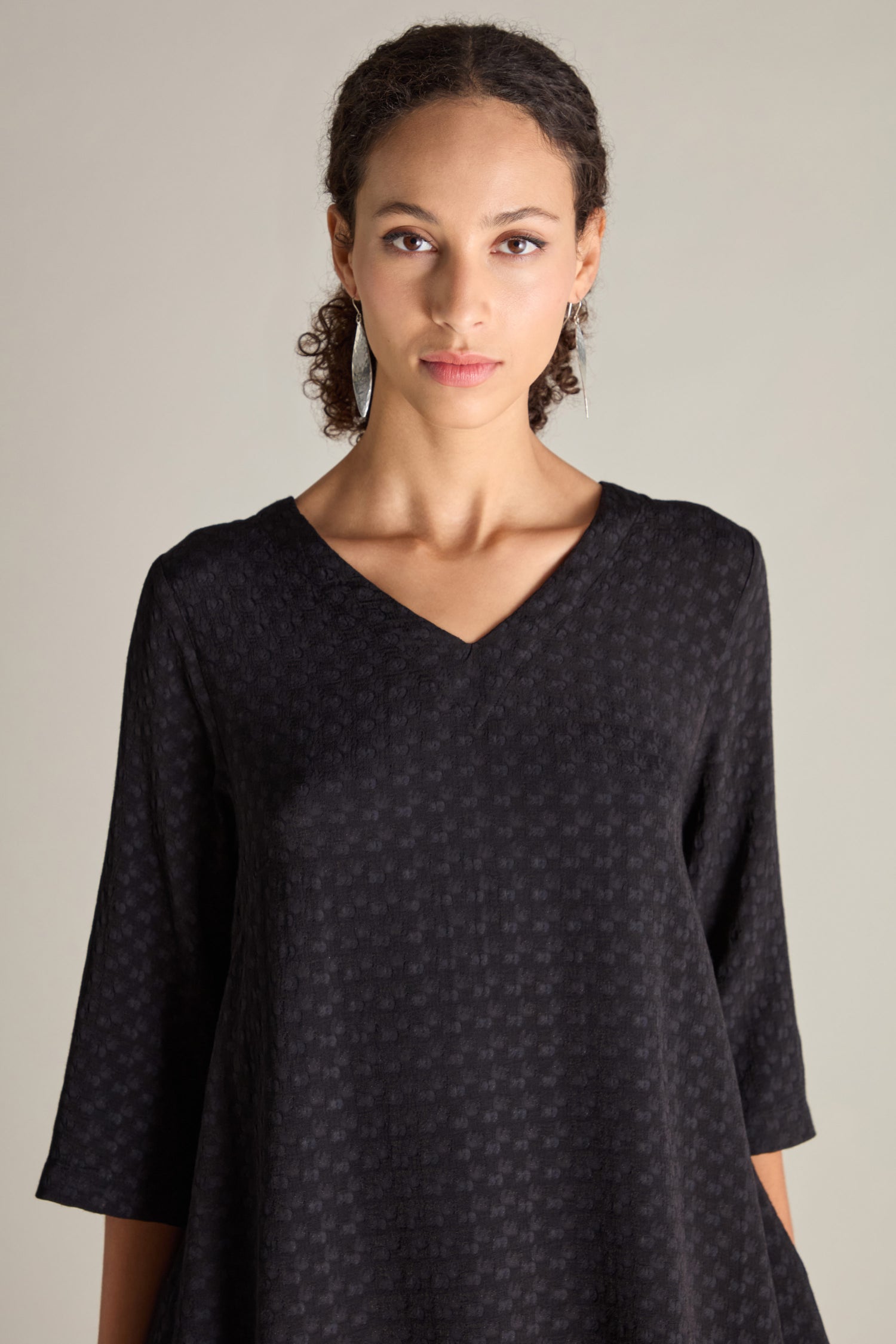 A person with curly hair is wearing a Small Square Jacquard Tunic with a black V-neckline and three-quarter sleeves, standing against a plain background.