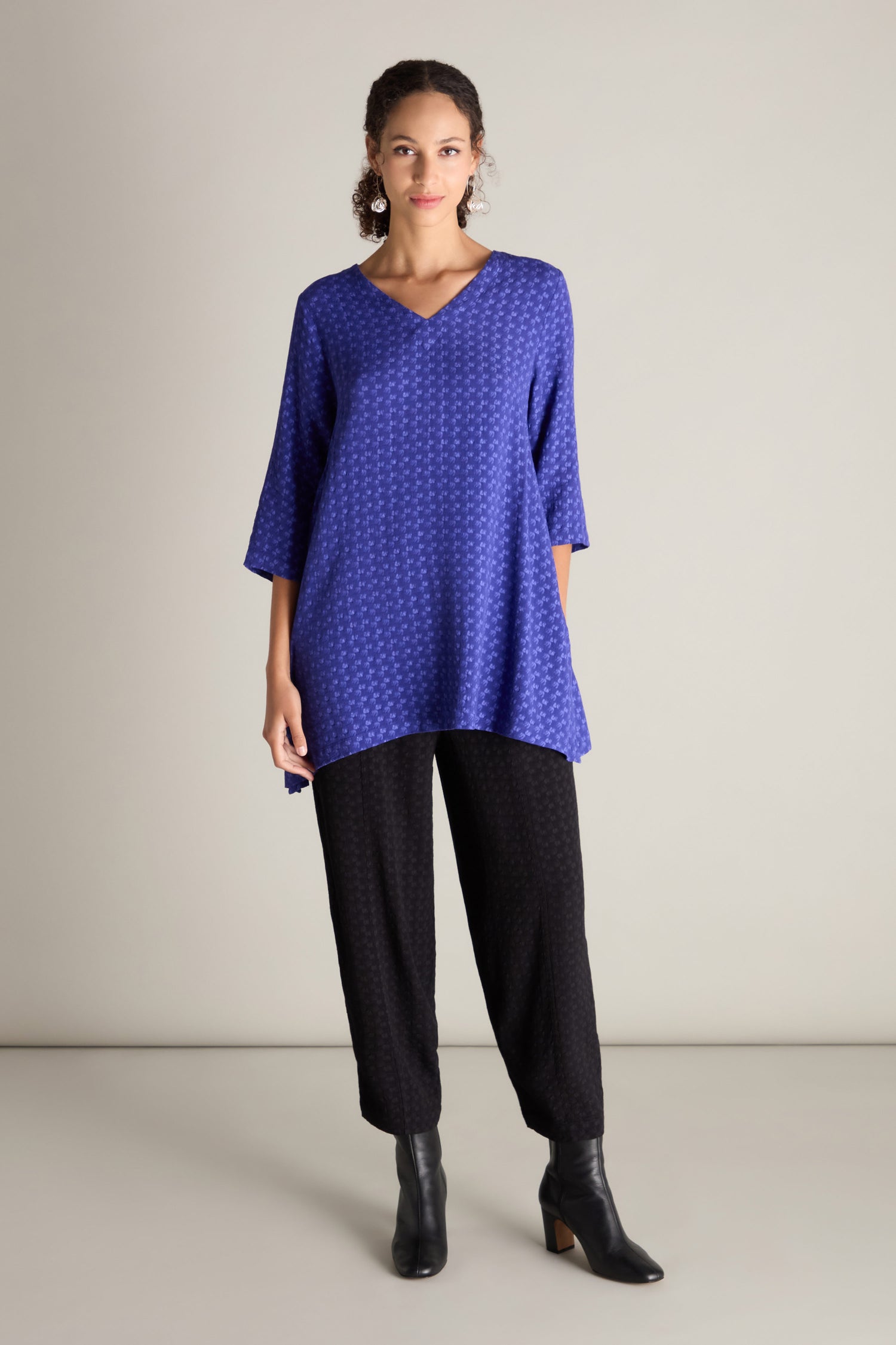 A person wearing a Small Square Jacquard Tunic, black pants, and black ankle boots stands against a neutral background. The tunic features 3/4 length sleeves and a delicate V-neckline, adding a touch of elegance to the ensemble.