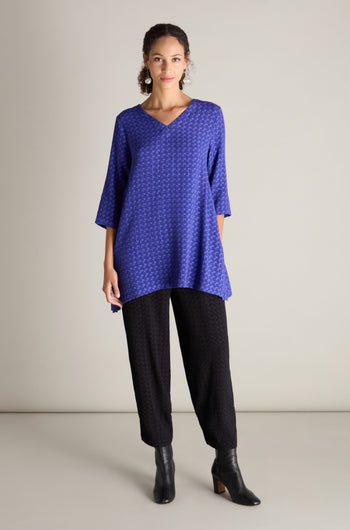 A person wearing a Small Square Jacquard Tunic, black pants, and black ankle boots stands against a neutral background. The tunic features 3/4 length sleeves and a delicate V-neckline, adding a touch of elegance to the ensemble.