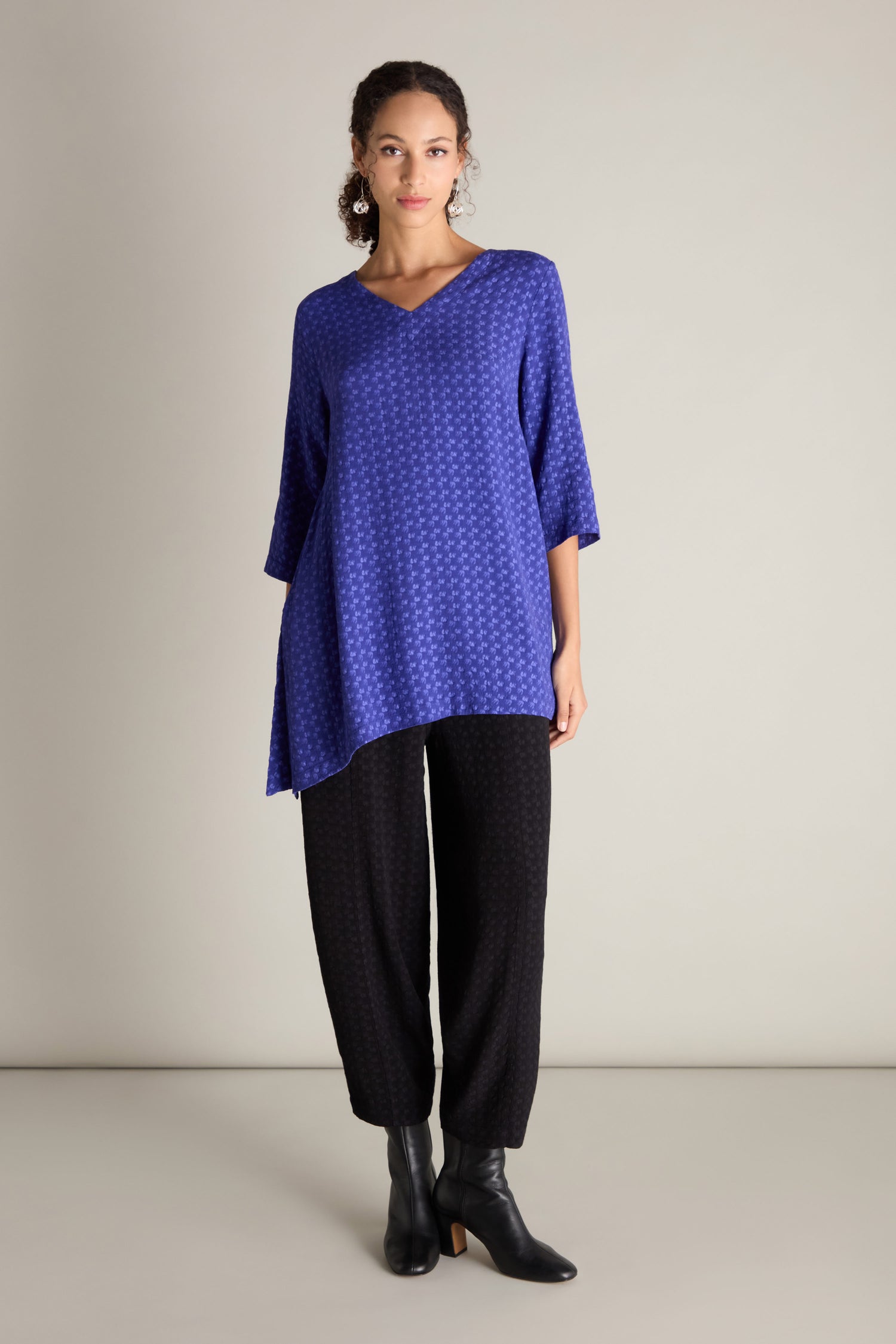 A person wearing a loose-fitting blue Small Square Jacquard Tunic featuring a V-neckline and 3/4 length sleeves, paired with black pants and black ankle boots, stands against a plain background.