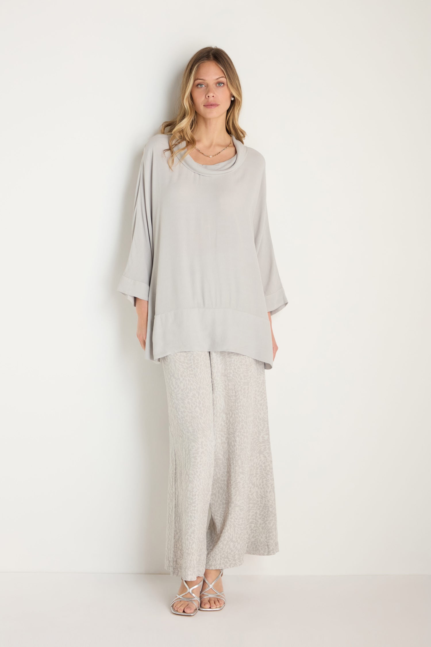 A woman stands against a white wall wearing a loose, light gray Crepe Boxy Top, wide-leg patterned light gray MTT5245-LVC pants, and strappy sandals.