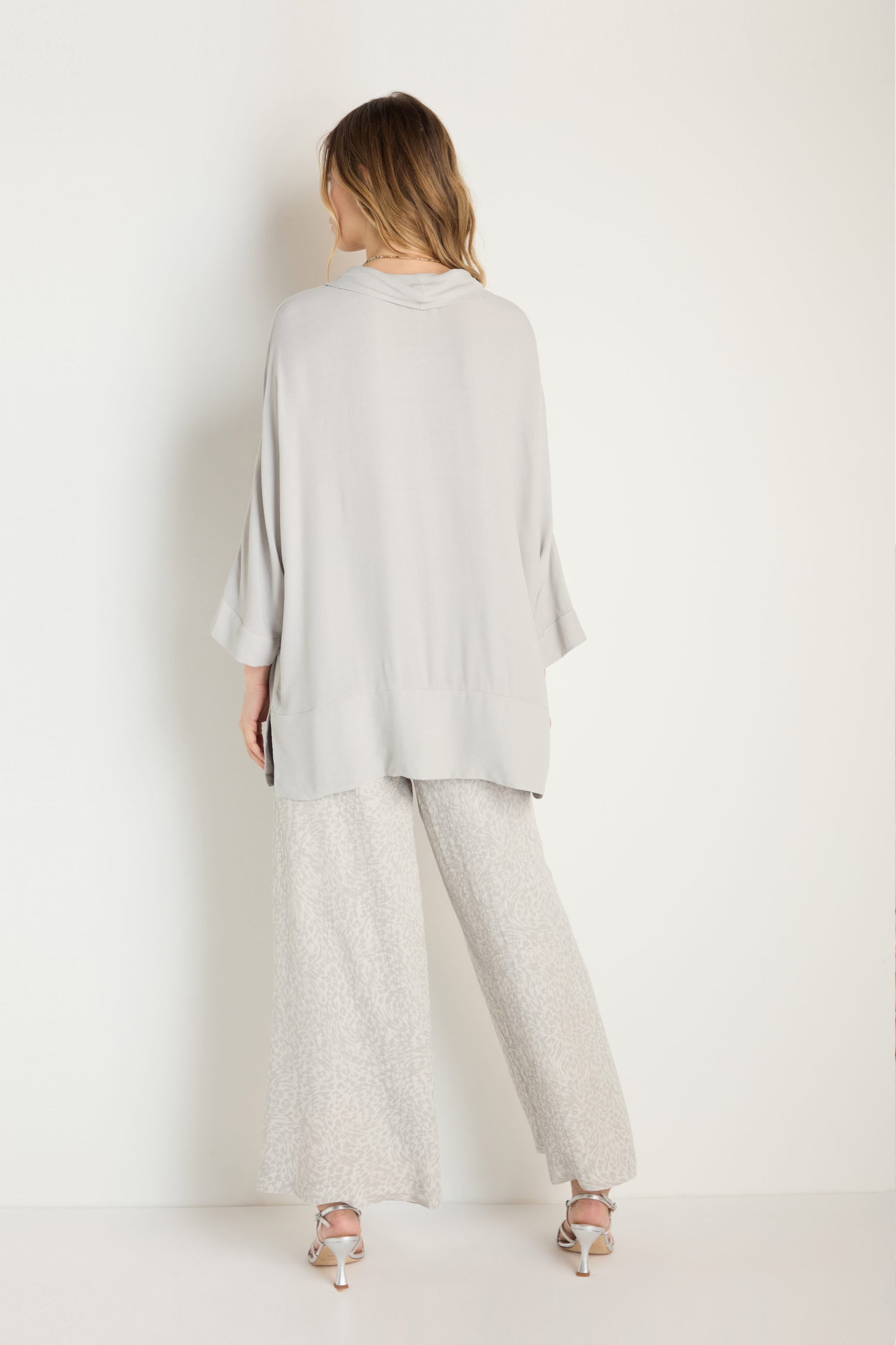 A woman stands facing a white wall, wearing a loose light gray Crepe Boxy Top, wide-leg patterned pants, and silver high-heeled sandals.