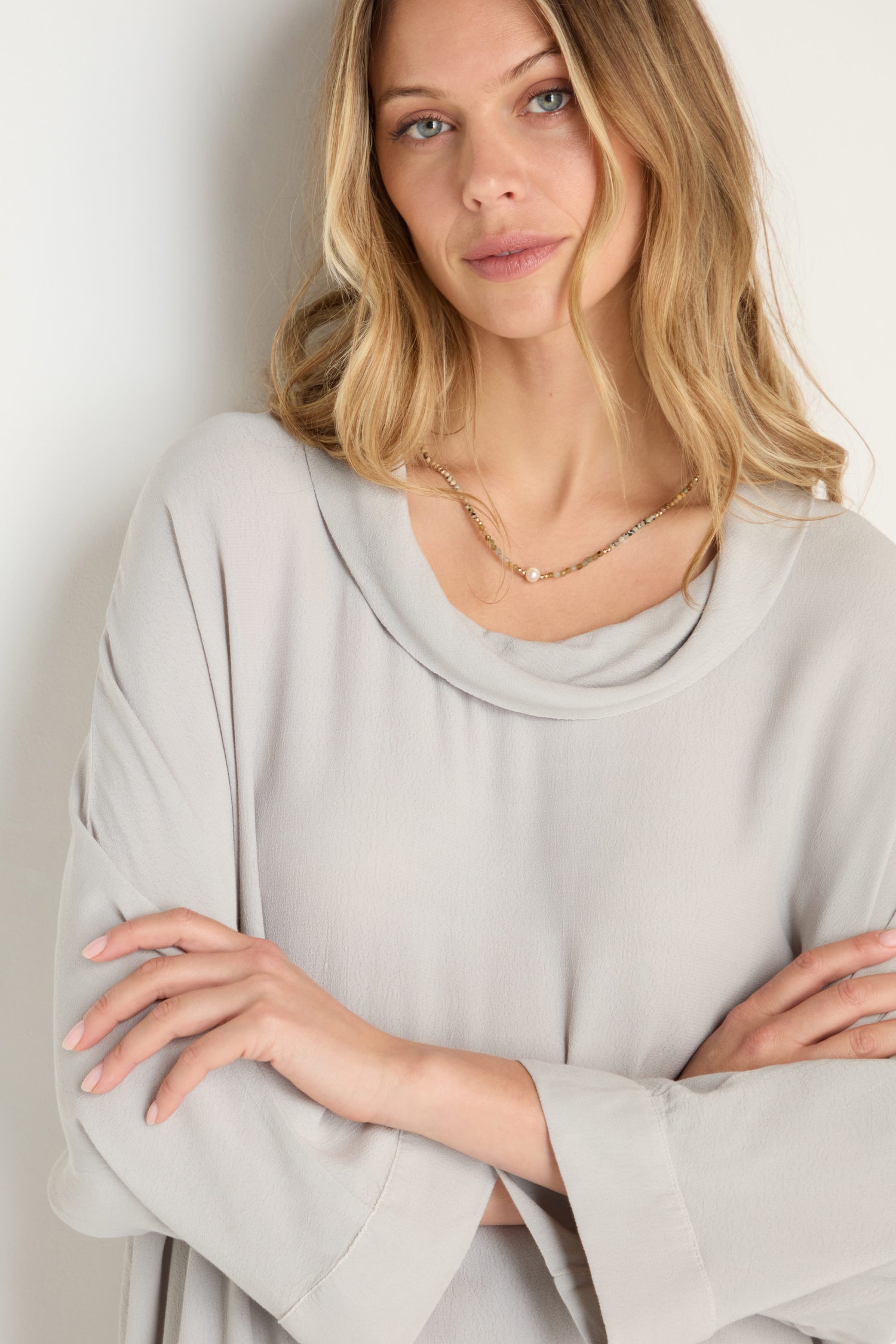 A woman with long blonde hair, wearing a loose, light gray Crepe Boxy Top and a gold necklace, stands with her arms crossed, looking at the camera.