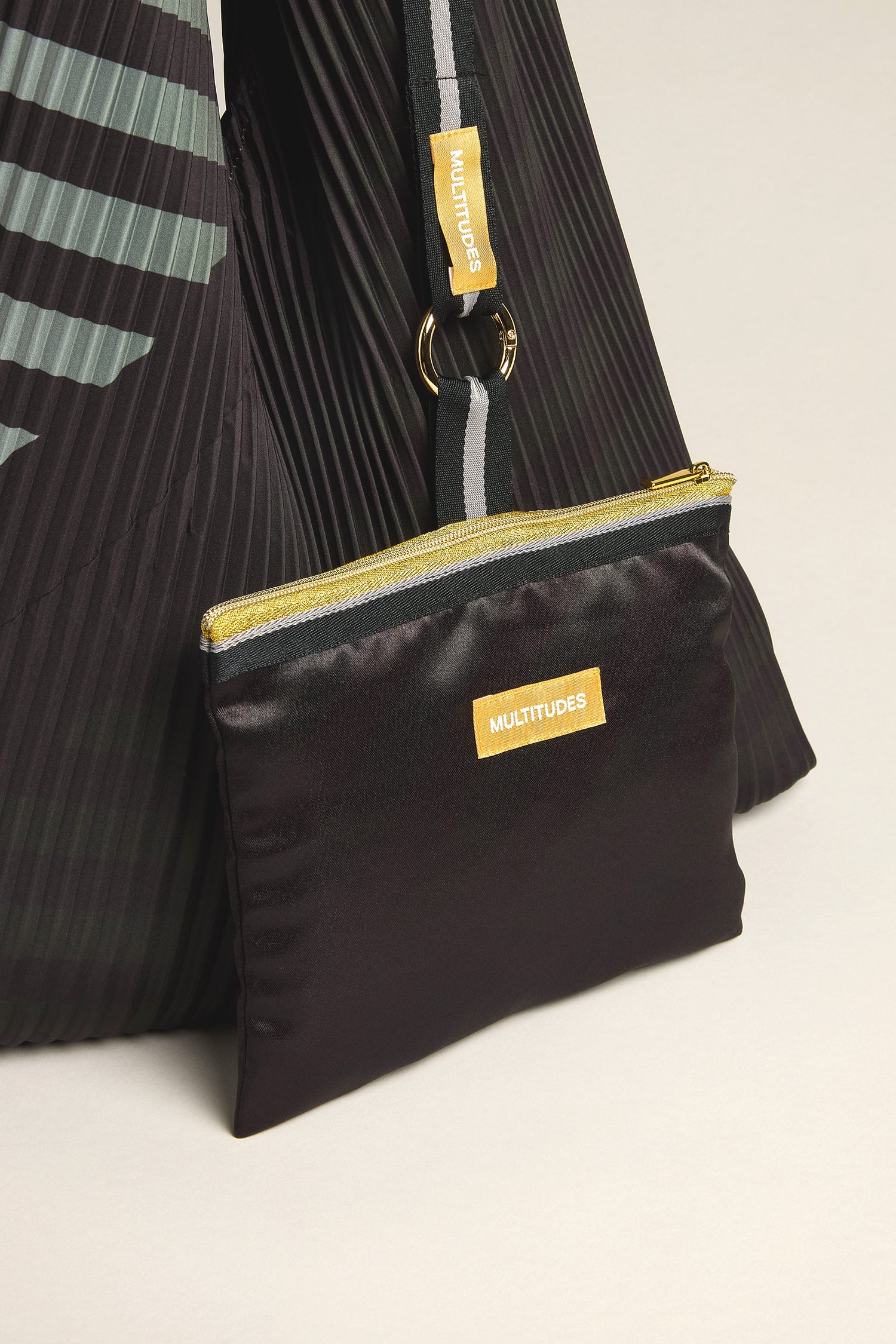 Close-up of the Pleated Y Handbag, featuring a black pouch with a yellow zipper and a tag labeled "MULTITUDES," attached to a striped strap, highlighting eco-friendly fashion through its sustainable design.