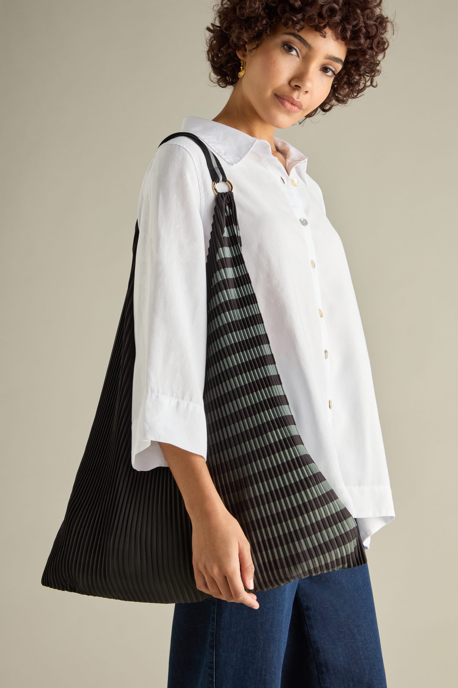 A person in a white shirt and jeans carries the Pleated Y Handbag, a large black pleated shoulder bag that highlights sustainable design with an eco-friendly flair.