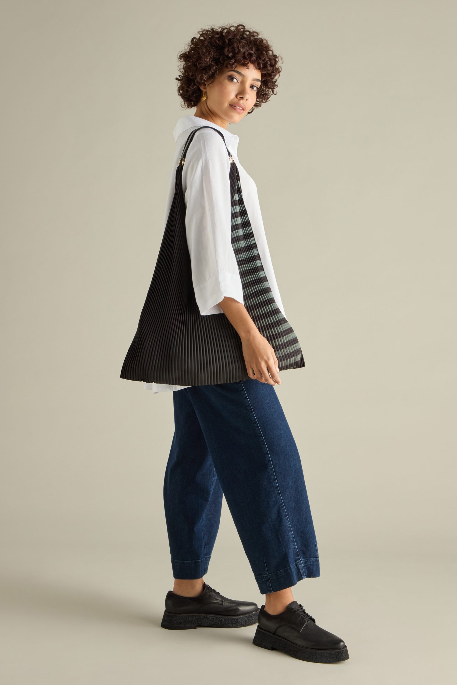 A person with curly hair in a white shirt and blue jeans poses with the eco-friendly Pleated Y Handbag, featuring a black striped design, facing slightly away from the camera.