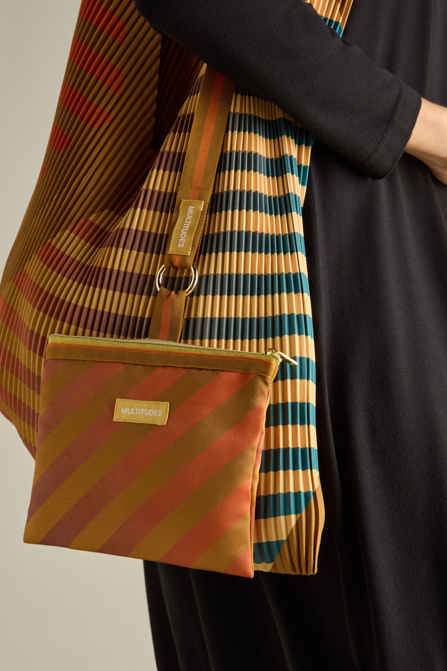 A person carries a sustainable Pleated Y Handbag with stripes in orange, yellow, and blue tones, paired with a black outfit.