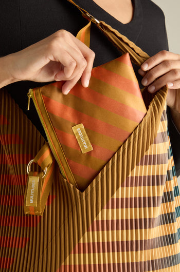 A person places a striped, multicolored zippered pouch into a Pleated Y Handbag, both labeled "Multitudes," highlighting eco-friendly fashion with their sustainable design.