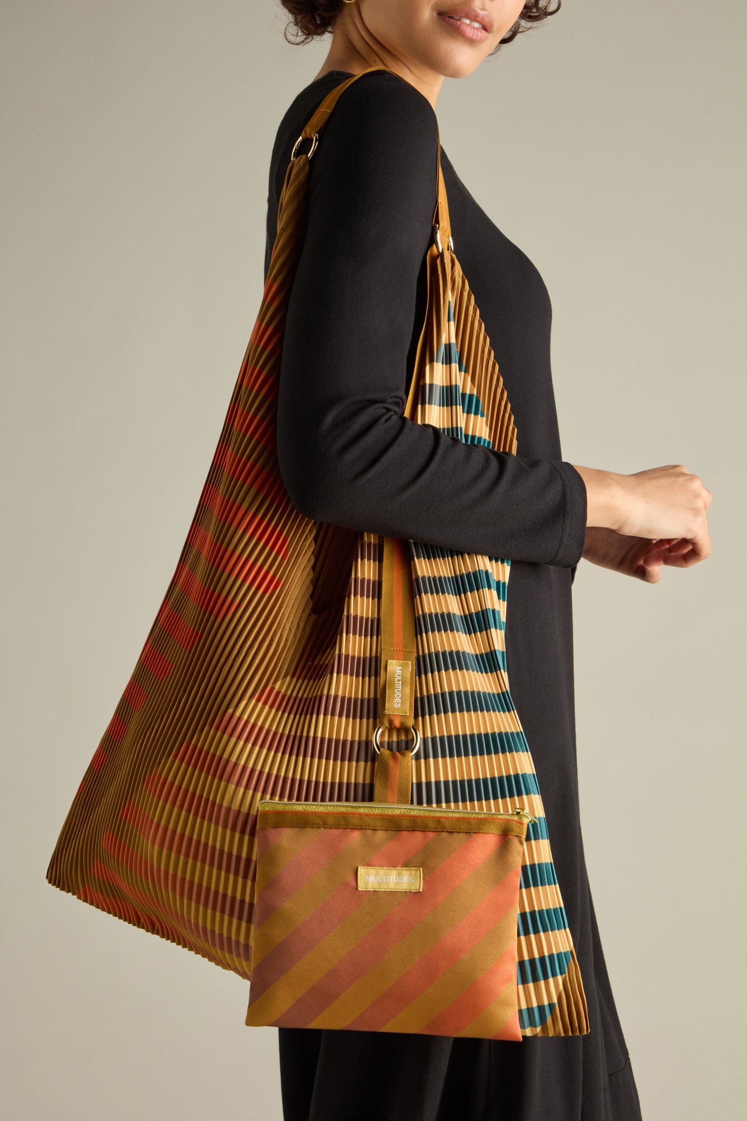 A person wearing a black dress carries an eco-friendly Pleated Y Handbag, a large striped recycled accessory with a matching smaller pouch in shades of gold, red, and blue.