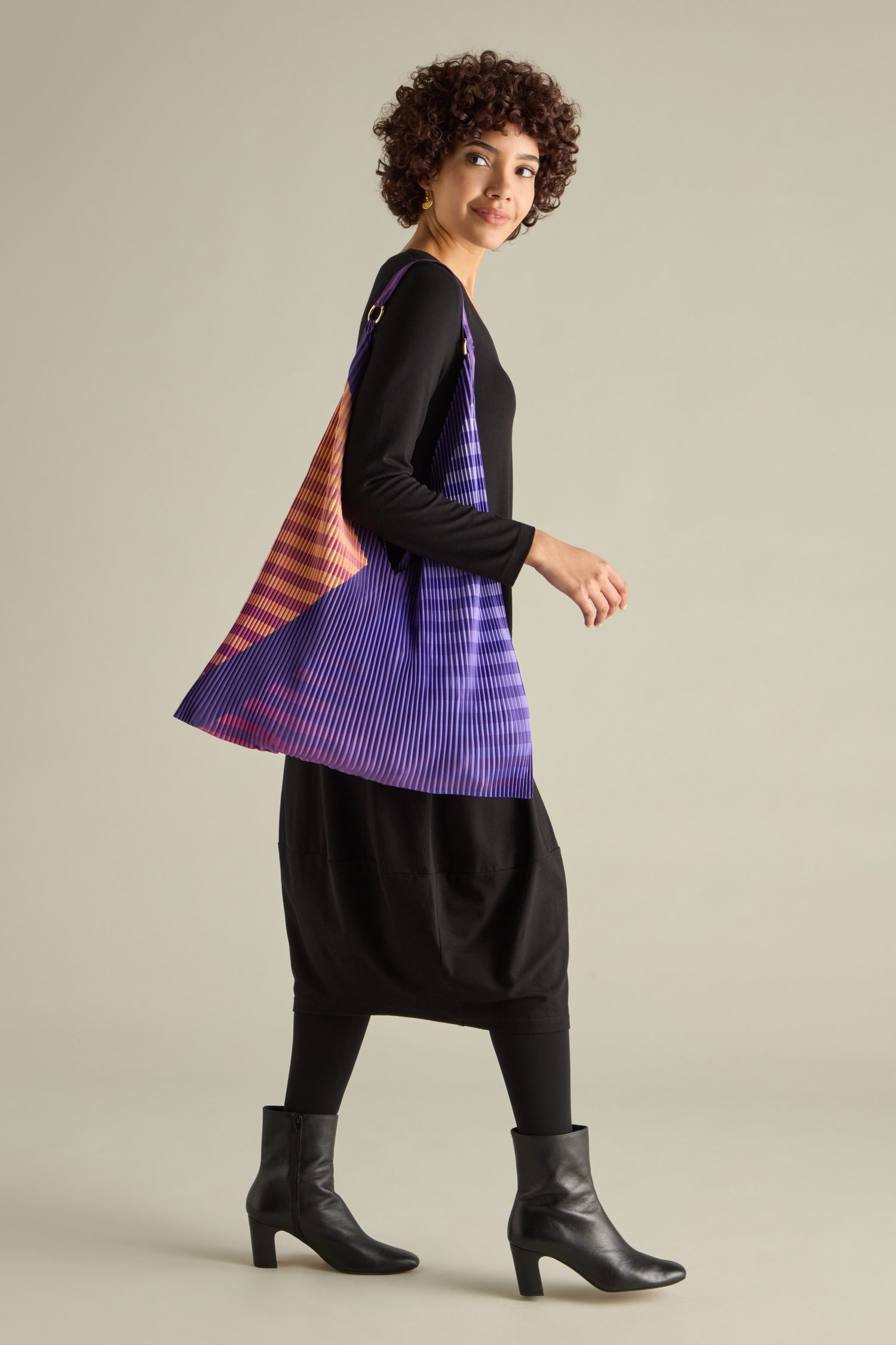 A person in a black dress and boots wears a colorful pleated shawl while gracefully carrying the Pleated Y Handbag indoors.