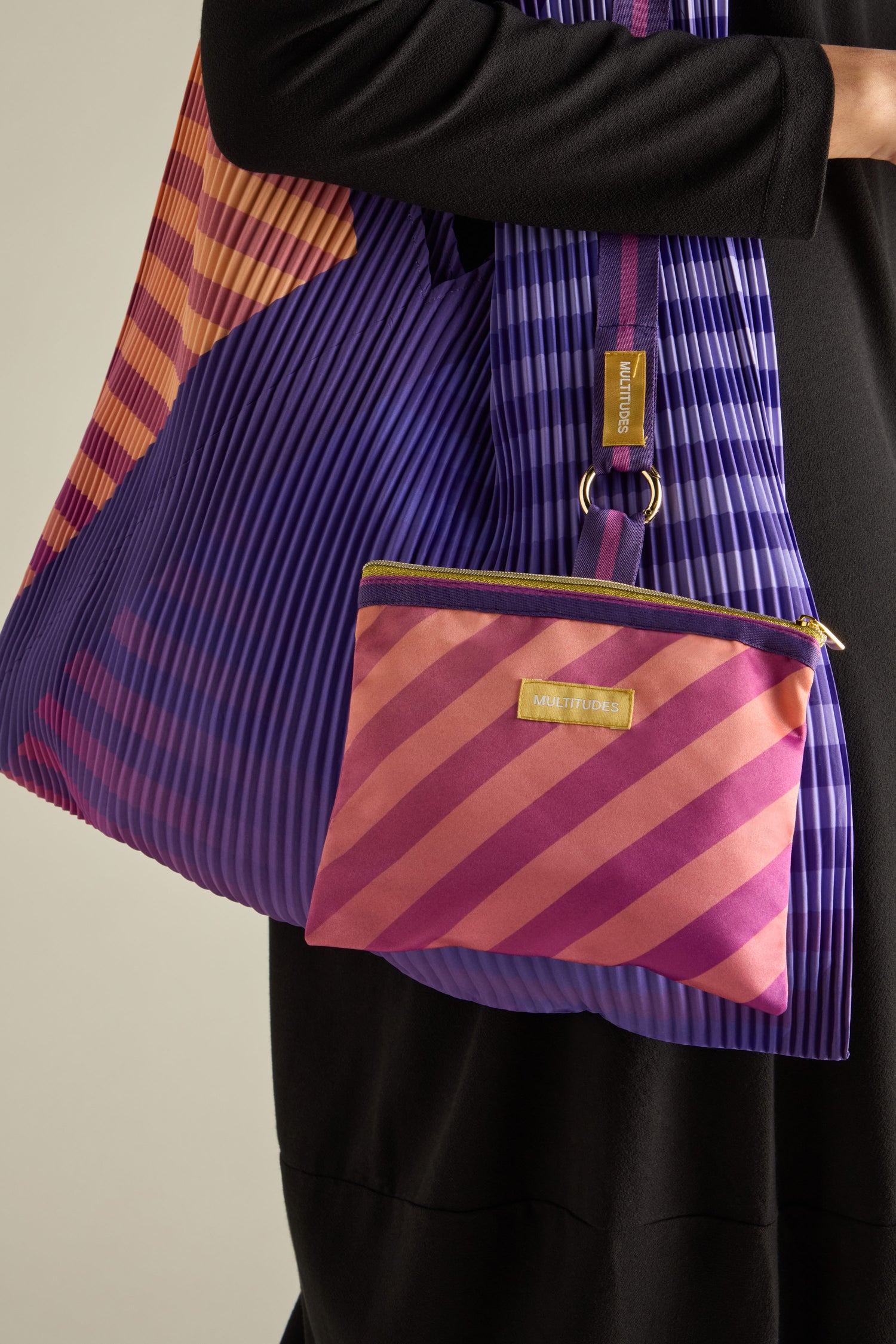 Someone is holding the Pleated Y Handbag, which is eco-friendly and displays a pleated purple design with orange and purple diagonal stripes, plus a matching small pouch.