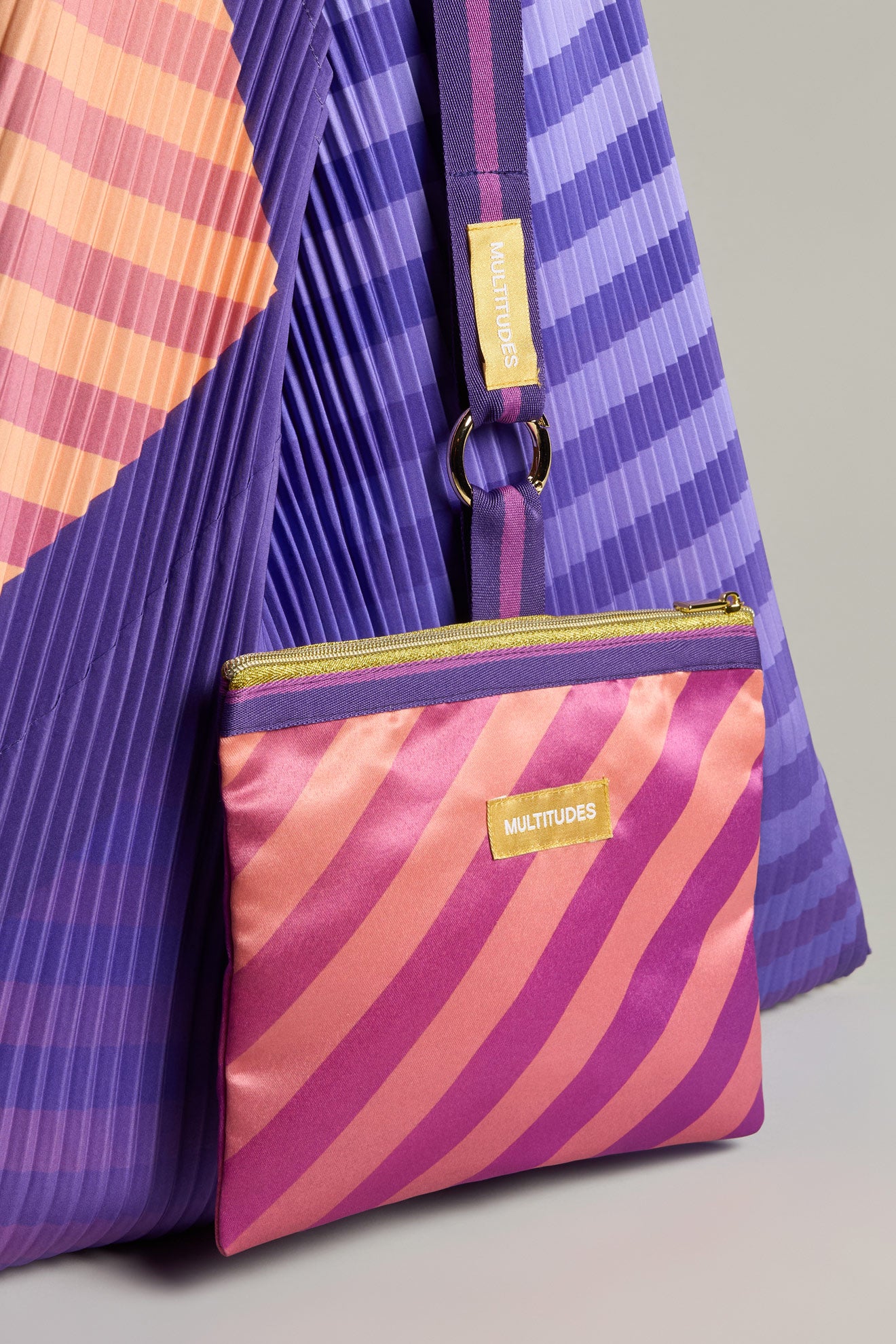 The "Pleated Y Handbag" is a recycled pouch with pink and orange stripes, featuring a gold "MULTITUDES" label, set against a purple and orange pleated fabric background.