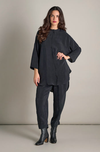 A person with long hair is wearing the Elba One Pocket Easy Cupro Shirt, a minimalist band-collar, loose-fitting black outfit, paired with ankle boots, standing against a plain background.