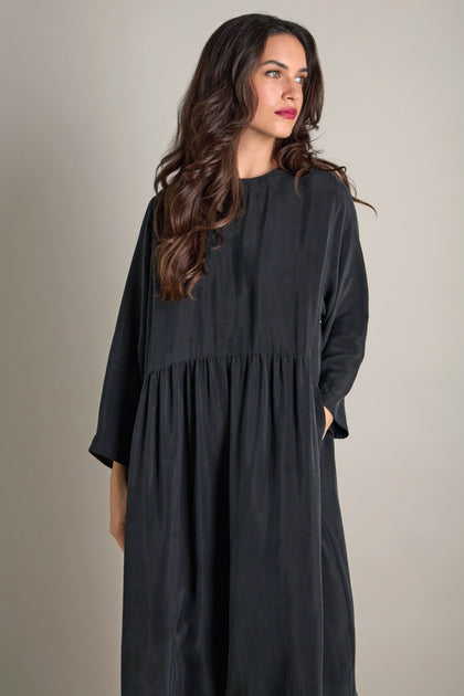 A person with long hair stands wearing a loose, dark Inga Cupro Drop Waist Dress against a plain background.