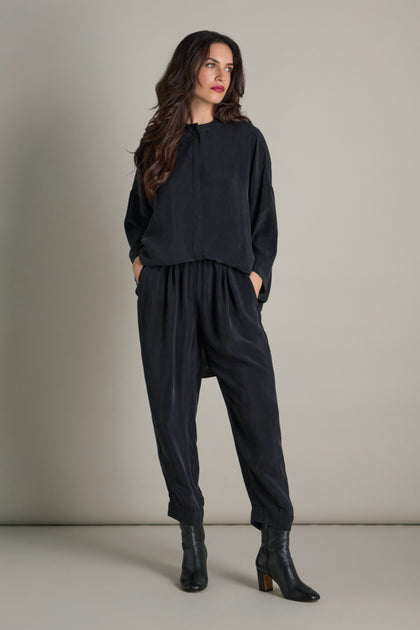 A person with long hair stands against a plain background, wearing a loose-fitting dark outfit that features the modern wardrobe staple, Lola Drapey Cupro Trouser with an elastic waistband. Their hands are tucked into pockets, complemented by black ankle boots.