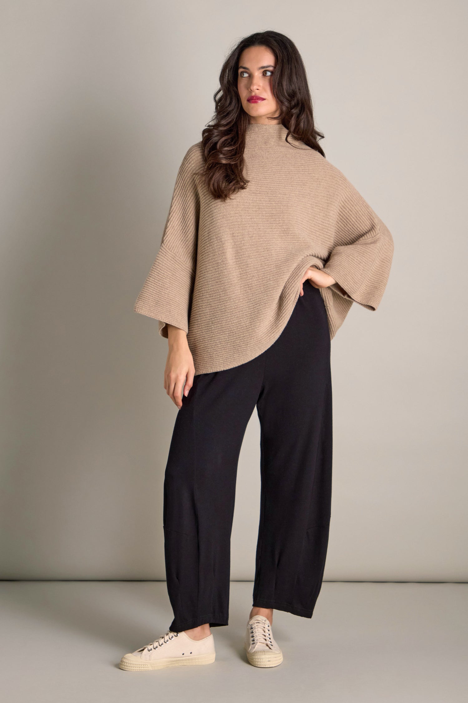 A woman stands against a plain background, wrapped in cosy comfort wearing the Pauline Rib Boxy Knit, an oversized beige sweater with a ribbed texture, paired with black pants and white sneakers.