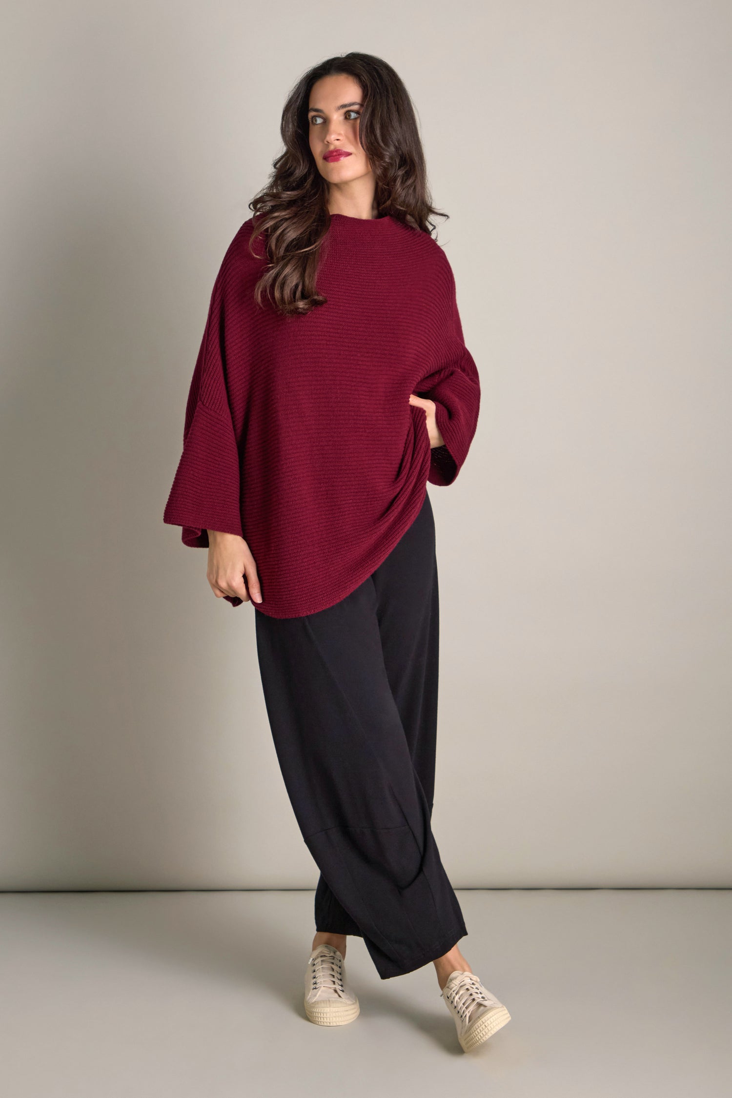 Against a plain background, a person models the Pauline Rib Boxy Knit—a maroon oversized sweater that exudes cosy comfort—paired with black pants and beige sneakers.