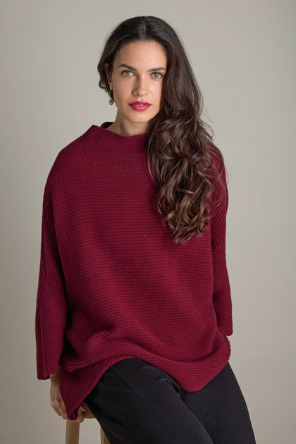 Person seated, wearing the luxurious Pauline Rib Boxy Knit in a cosy burgundy hue, paired with black pants, long brown hair cascading against a neutral background.