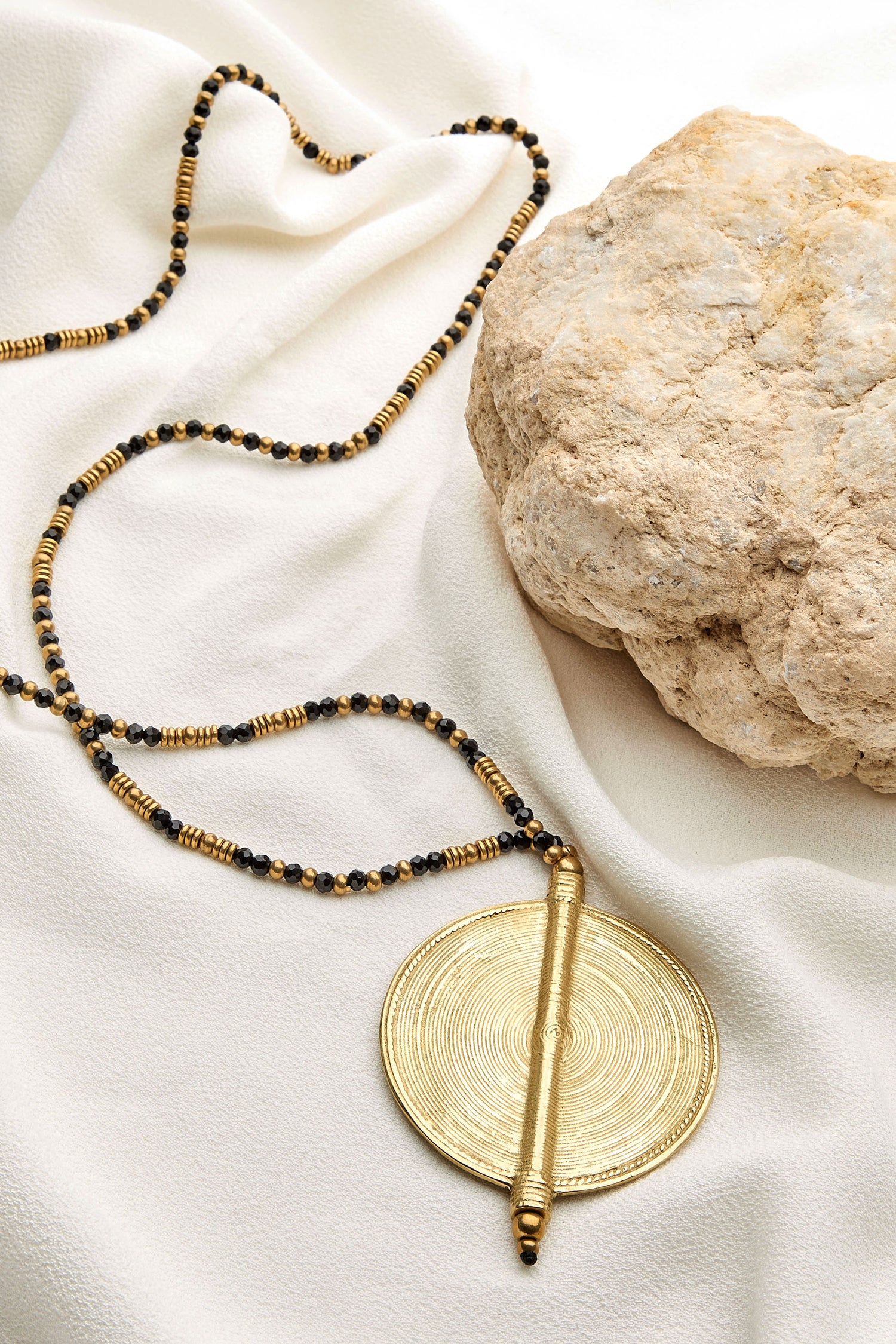 The Spiral Pendant, a long gold and black beaded necklace with a large round gold charm, is elegantly placed on white fabric near a textured beige rock.