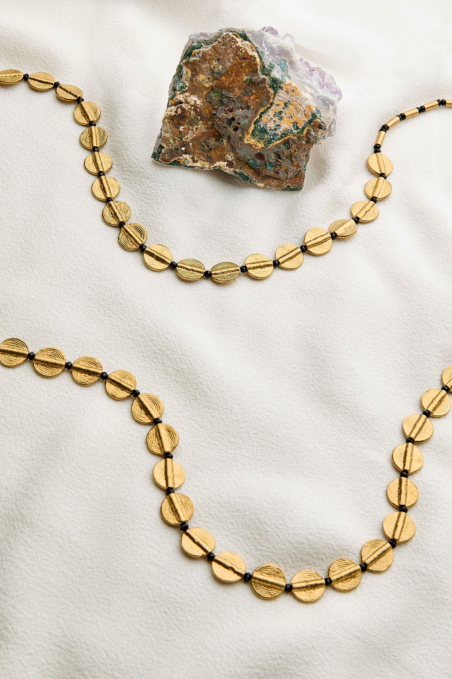 An Engraved Circles Necklace is displayed on a cream surface, next to a multicolored piece of rough mineral. Meticulously crafted by Jaipur artisans, this gold necklace combines tradition with elegance.