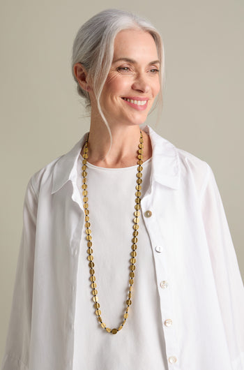 A woman with gray hair, wearing a white blouse and the Engraved Circles Necklace crafted by Jaipur artisans, smiles while looking to her right.