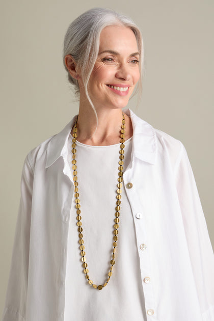 A woman with gray hair, wearing a white blouse and the Engraved Circles Necklace crafted by Jaipur artisans, smiles while looking to her right.