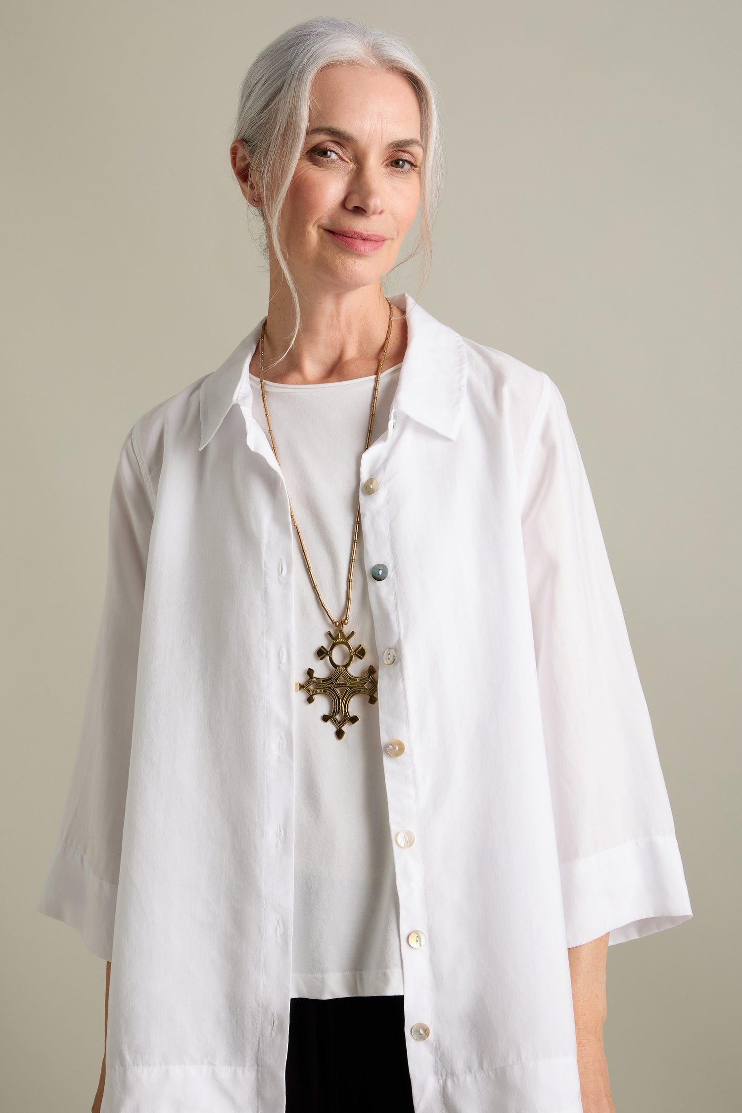 A woman with gray hair tied back, wearing a white blouse and a Bartchakea Tuareg Pendant adorned with gold bugle beads, stands against a plain background.