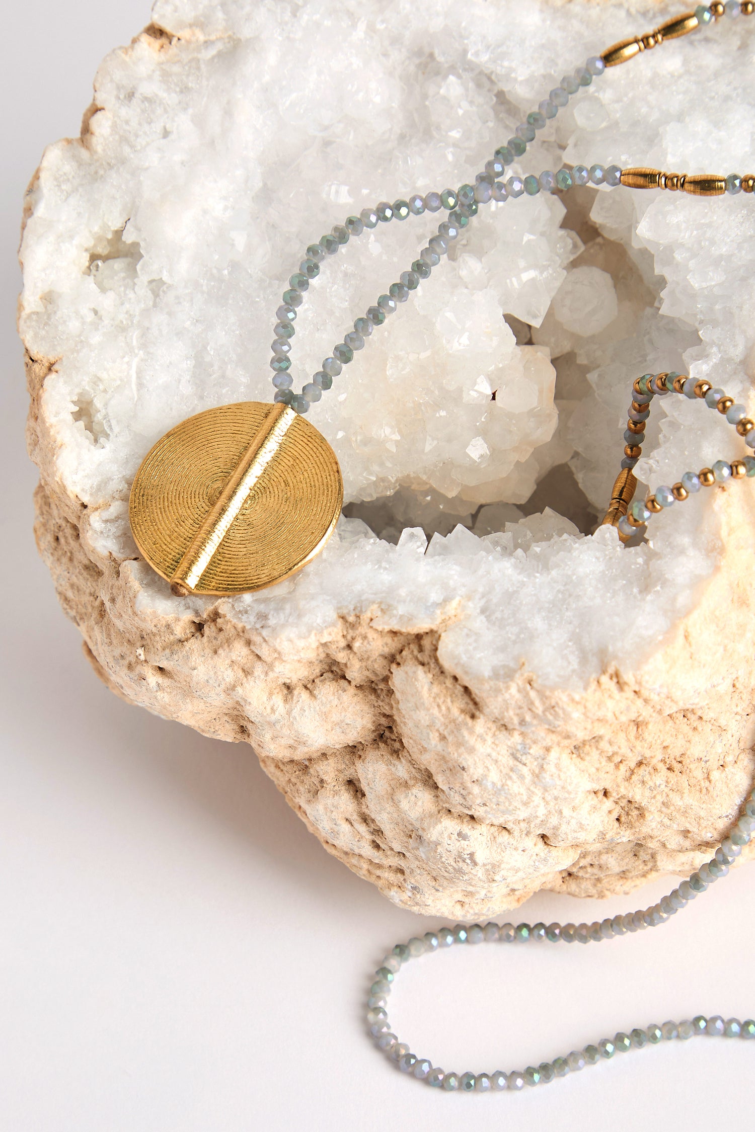 The Moana Spiral Bead Pendant, featuring elegant gold and small gray beads, is artfully draped over a large rough geode with a crystalline interior, highlighting exquisite artisan craftsmanship.