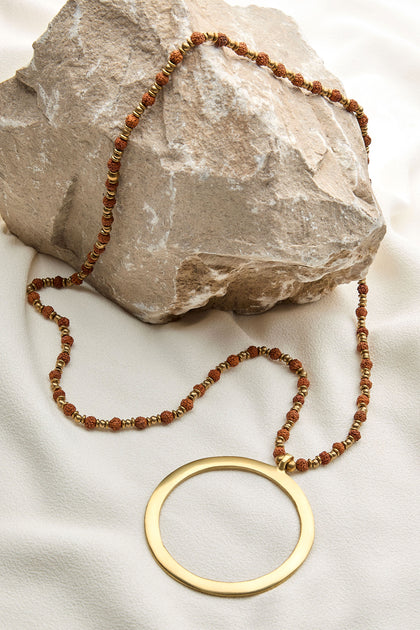 Circle Pendant with a large gold ring pendant and brown beaded chain, featuring intricate Rudraksha seeds, displayed draped over a rock on a light fabric background.