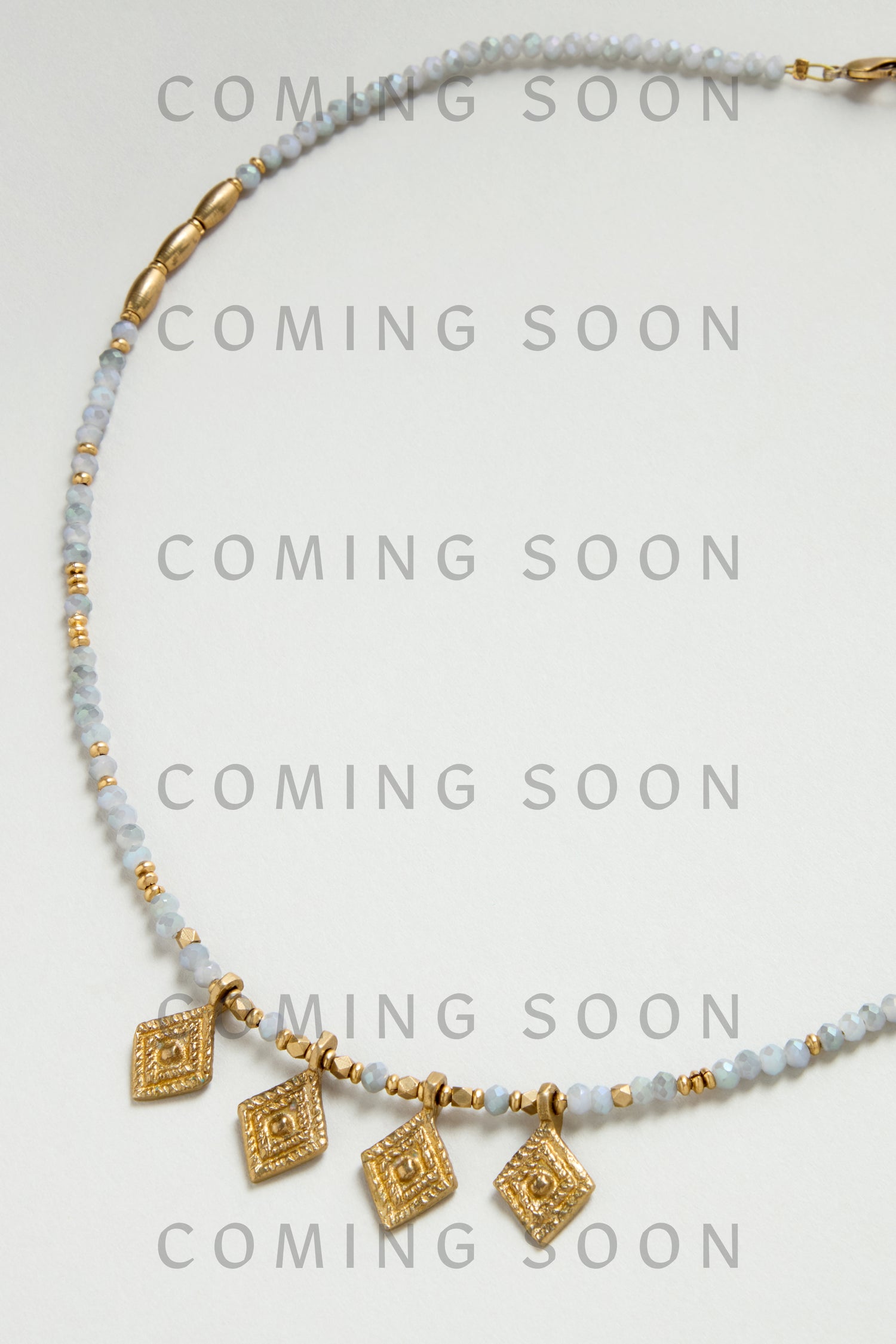 The Eanna Necklace features gold geometric charms with small blue and gold beads, shown on a light background with "COMING SOON" text. Style code: NCCCE363.
