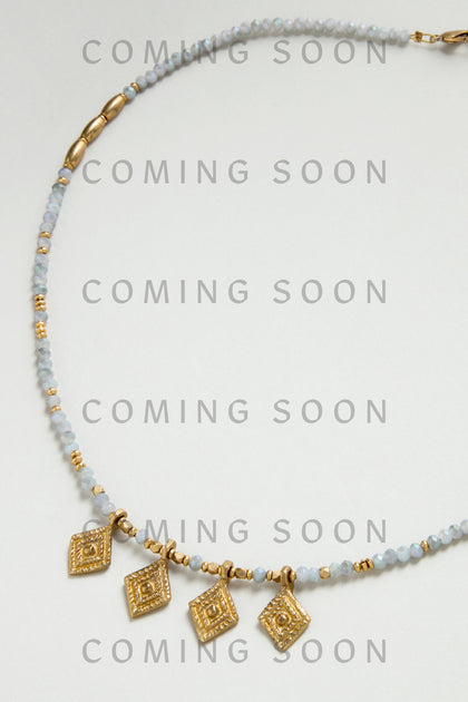 The Eanna Necklace features gold geometric charms with small blue and gold beads, shown on a light background with "COMING SOON" text. Style code: NCCCE363.