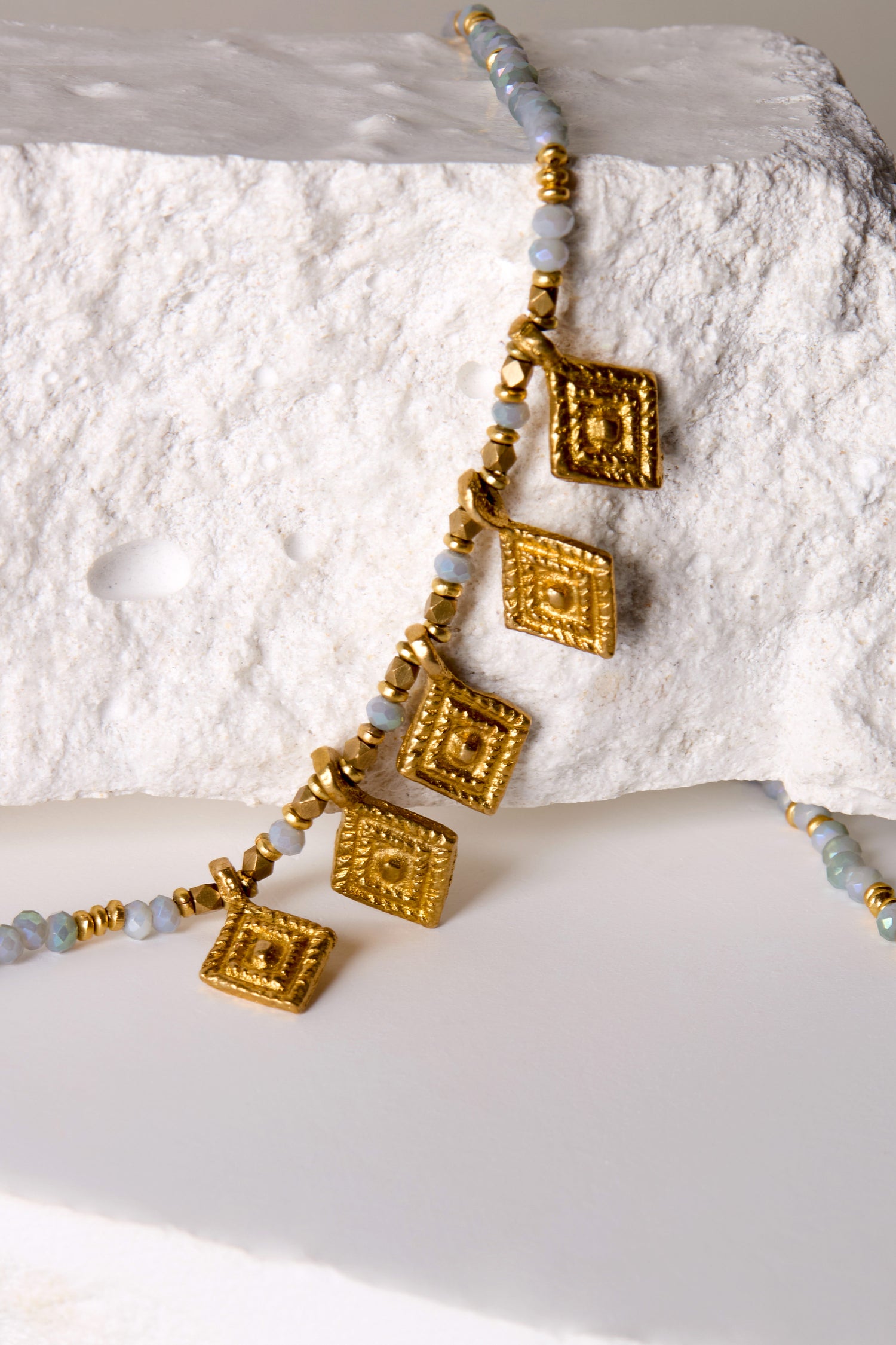 The Eanna Bead Pendant necklace showcases gold-tone beads and four elegant square pendants, all beautifully arranged on a textured white background.