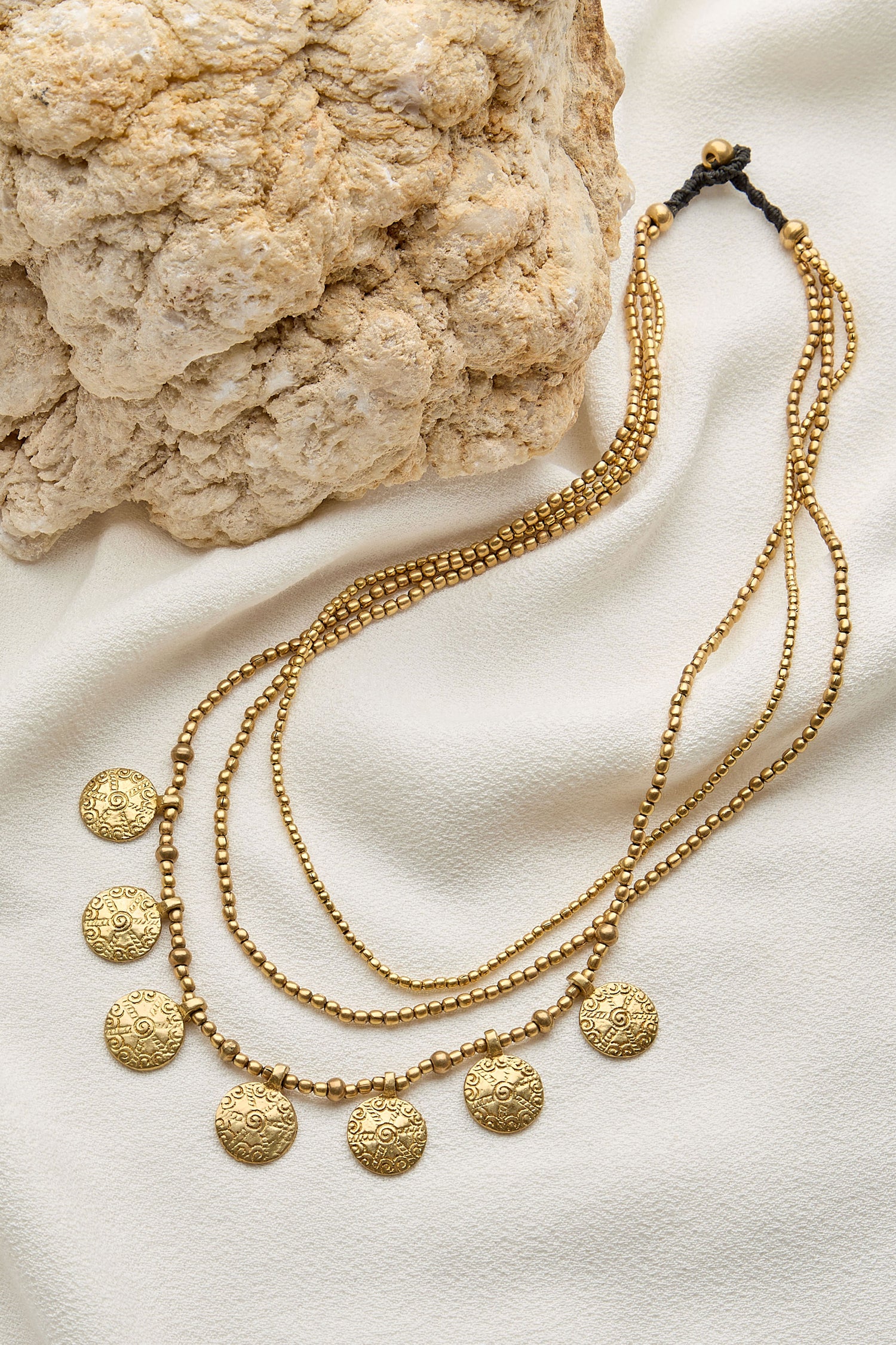 A Chakra Necklace featuring multiple layers of gold beads and round engraved chakra pendants, displayed on cream-colored fabric next to a textured rock.