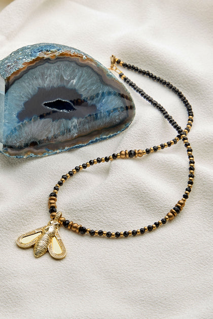 A Bumblebee Necklace, adorned with gold and black beads and featuring a decorative bumblebee pendant, is displayed on a light fabric beside a sliced geode.