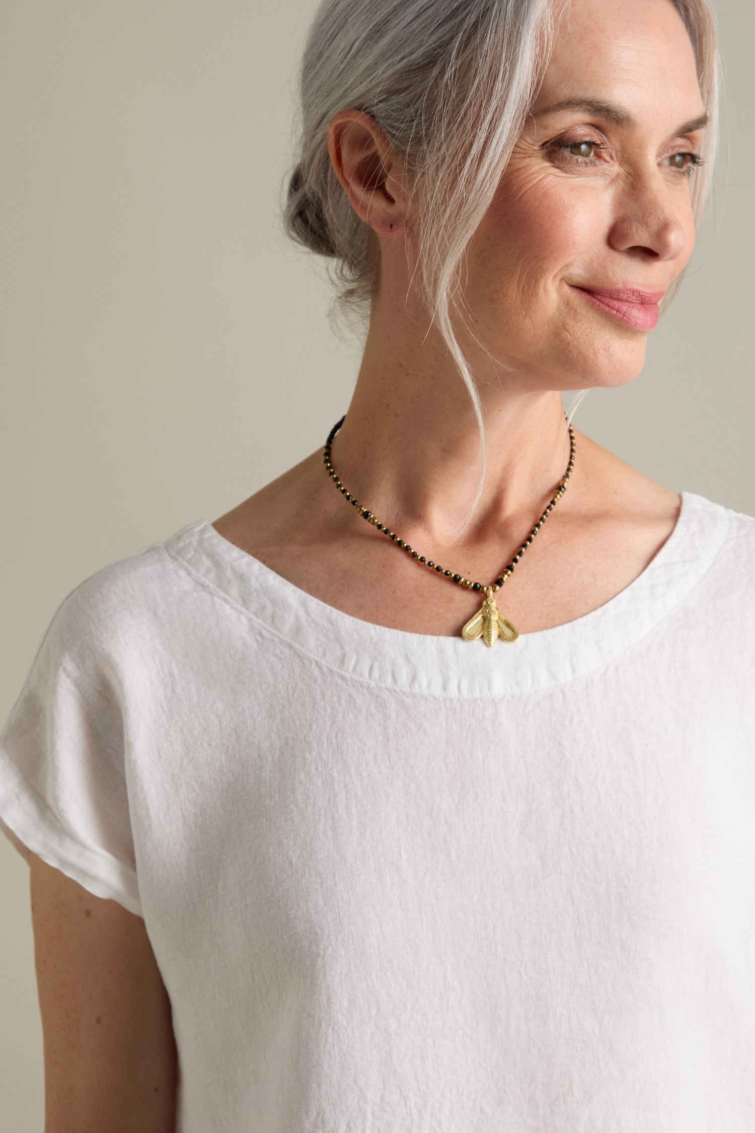 An older woman with grey hair stands smiling, wearing a white blouse and a Bumblebee Necklace adorned with gold beads.